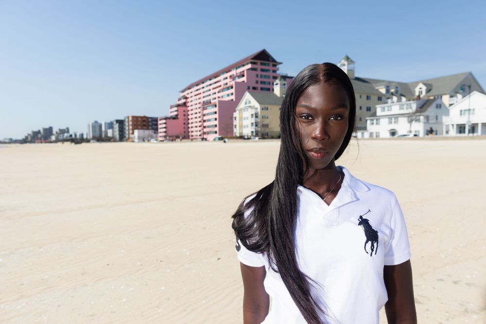 Young Black woman in white Ralph Lauren polo stands on sandy beach with colorful buildings in background.