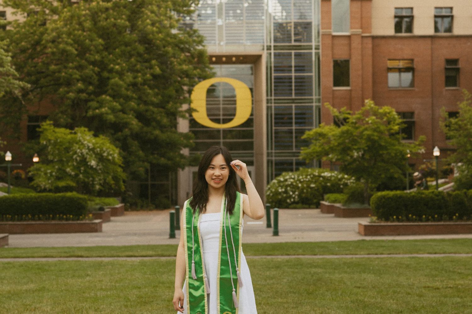 A graduate in green graduation stoles stands smiling on a lawn with the University of Oregon O logo in background. Oregon senior photos