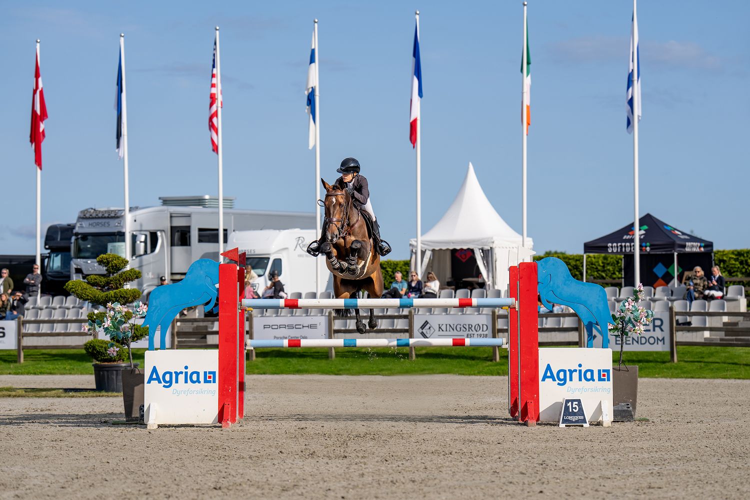 Horse and rider clearing a jump at competition with Norwegian flag flying overhead.