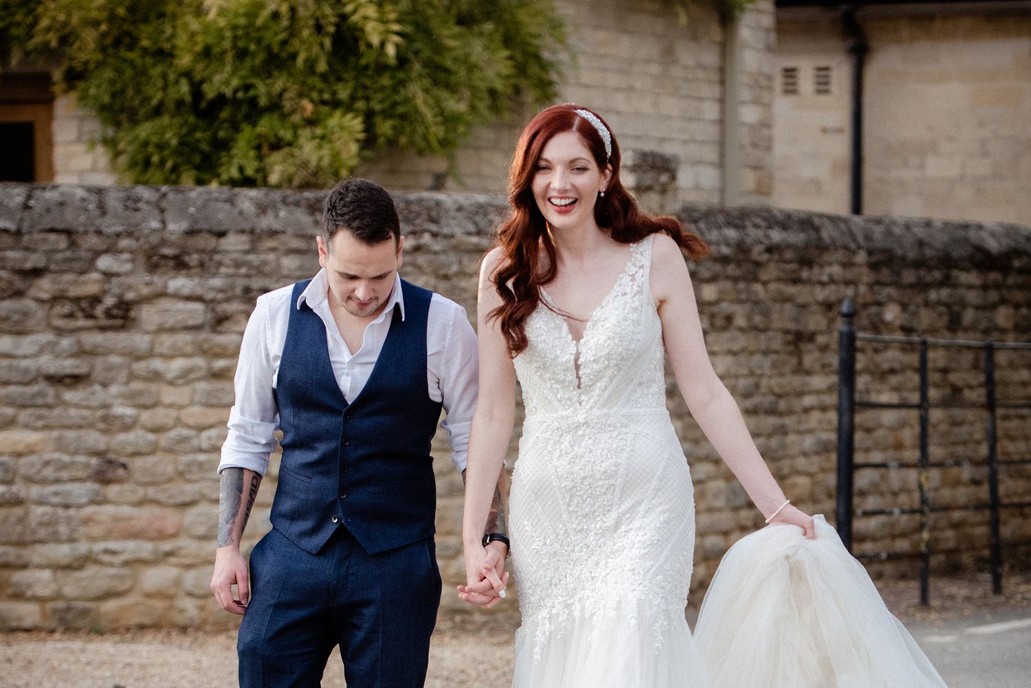 Couple sharing joyful moment while walking beside stone wall in wedding attire.