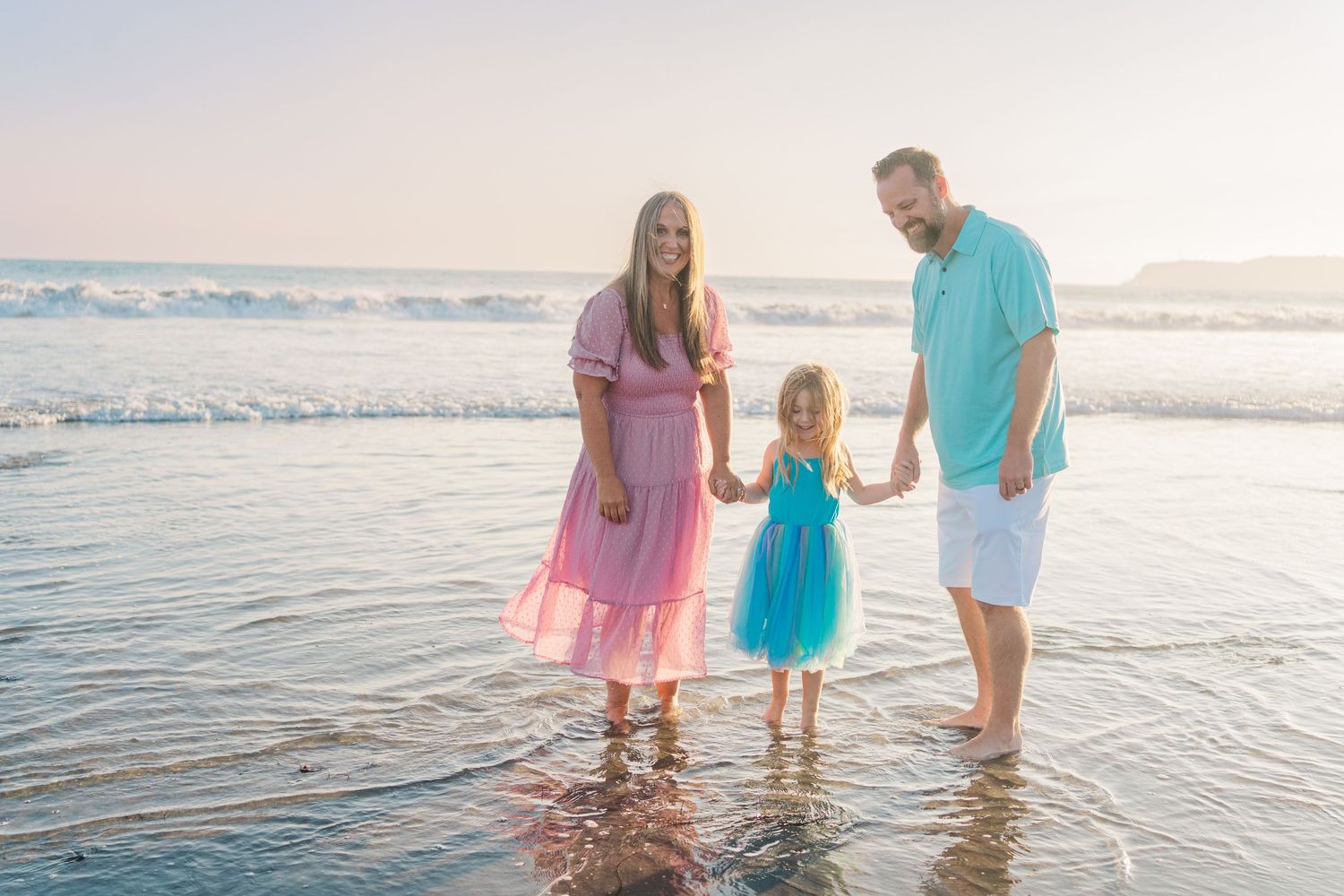 A family stands together in shallow ocean waves at sunset while wearing pink, turquoise and white outfits.