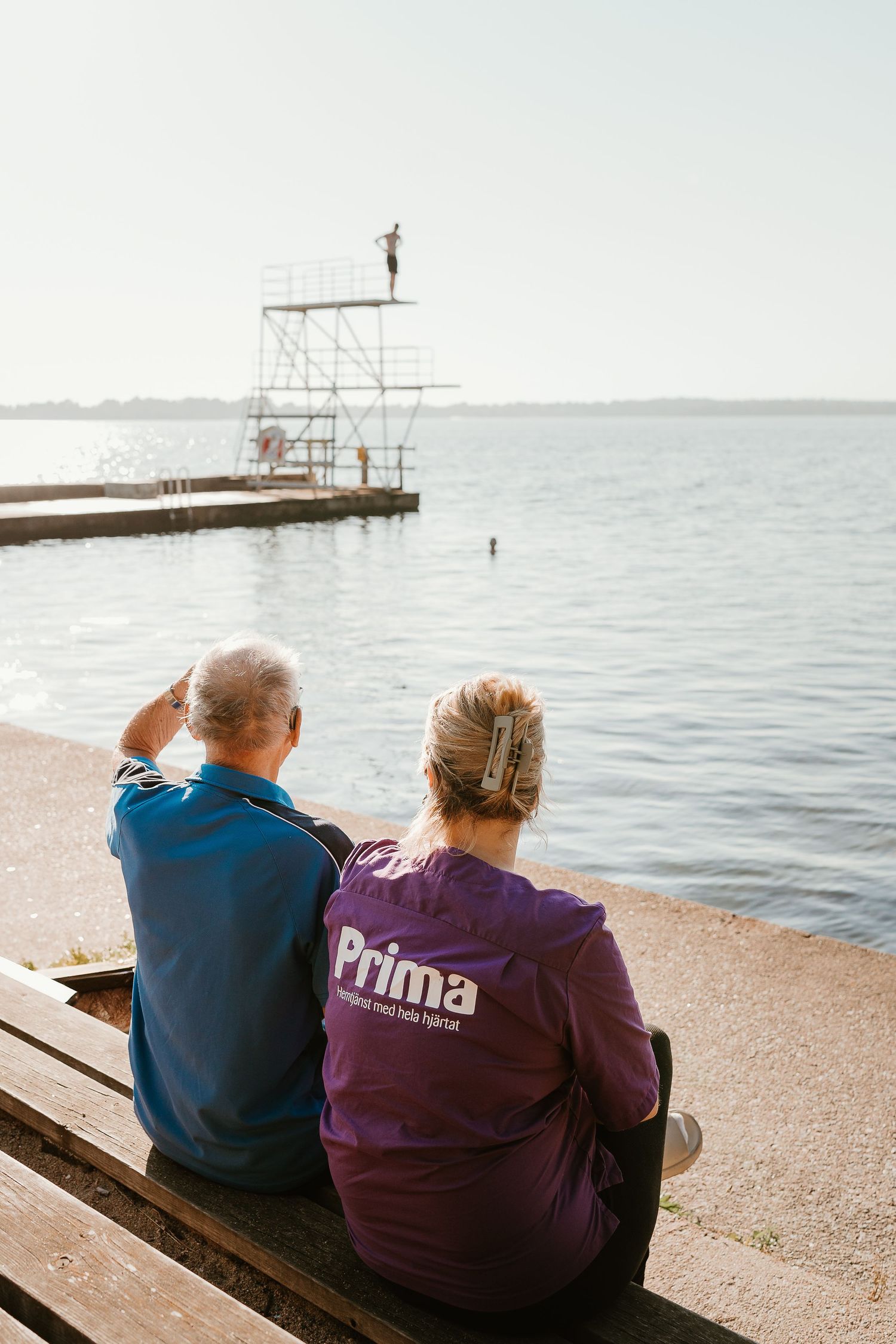 Two people sit on a bench overlooking a calm sea with a diving platform in the distance.