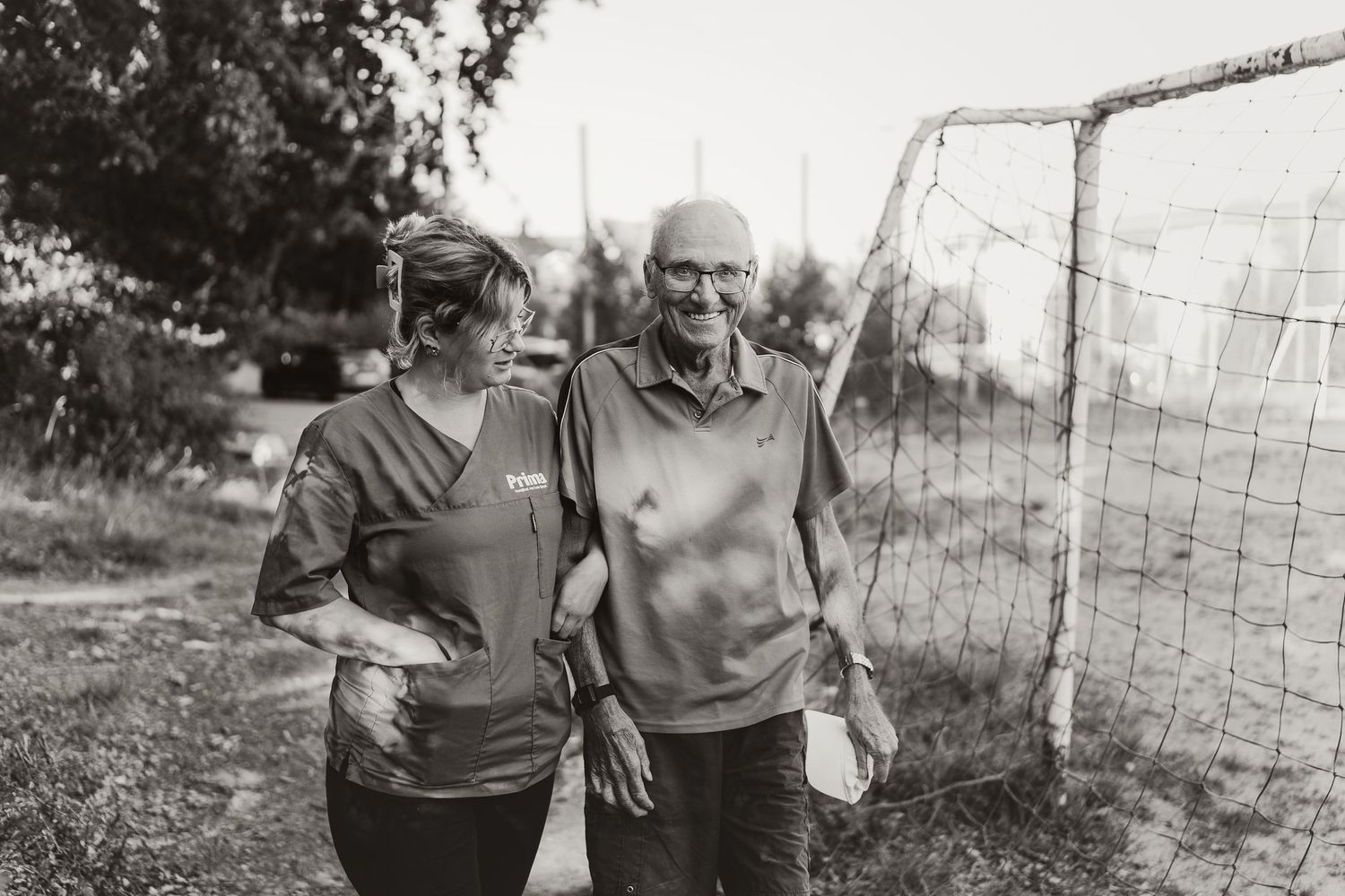 A caregiver and elderly person share a warm moment together near a soccer goal.