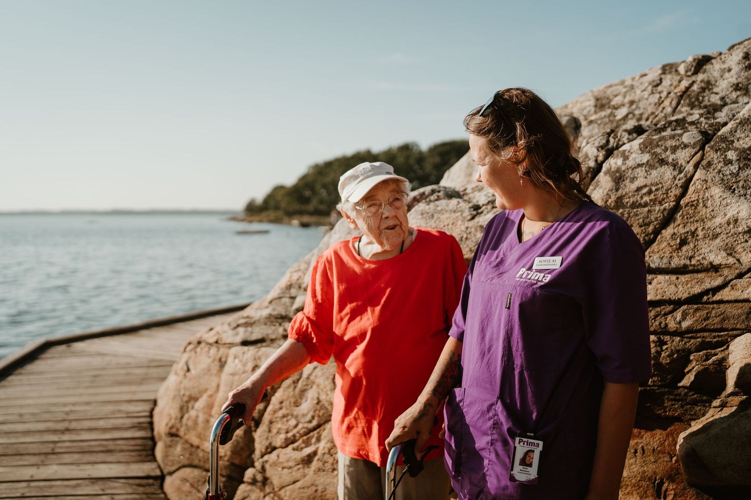 Healthcare workers in purple scrubs and red jacket sharing a moment on wooden boardwalk by rocky coastline.
