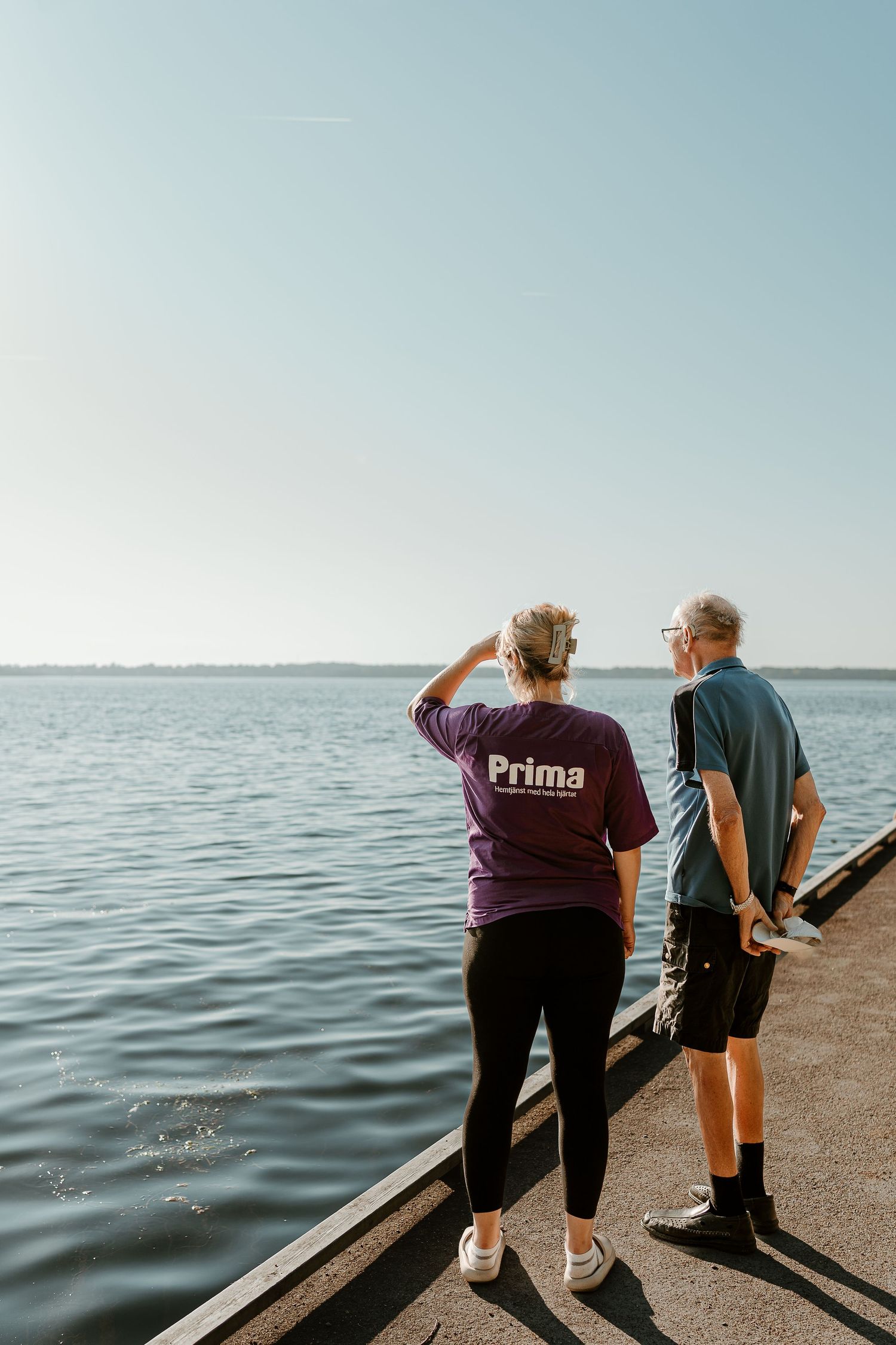 Two people looking out at calm waters from a pier while wearing Prima branded shirts.