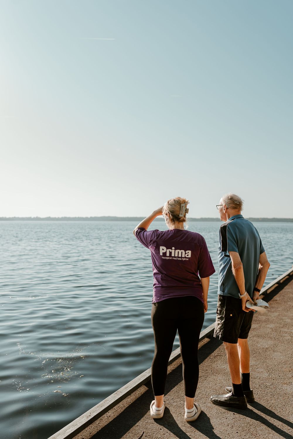 Two people looking out at calm waters from a pier while wearing Prima branded shirts.
