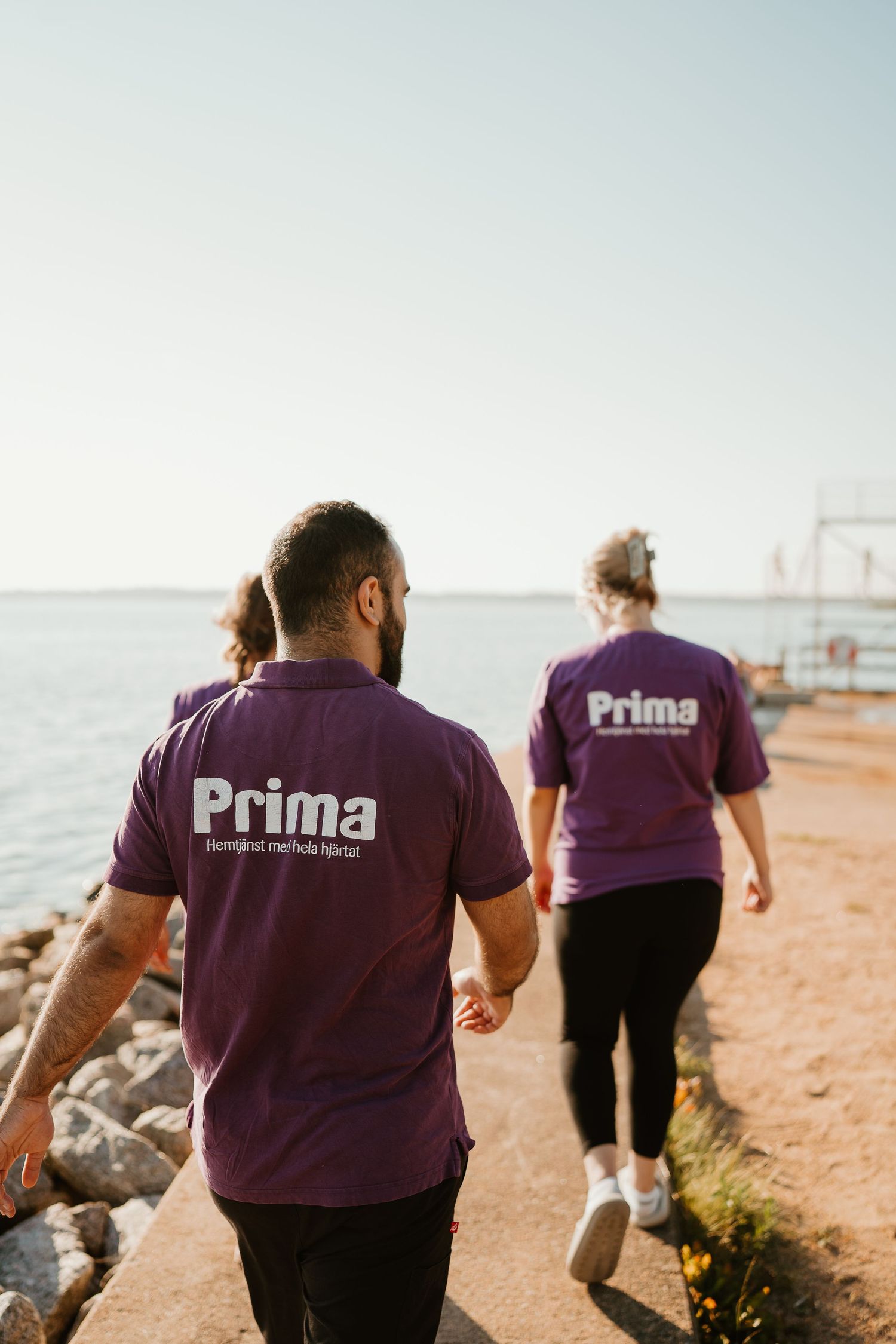 Staff members in purple Prima uniforms walking along coastal path near water.