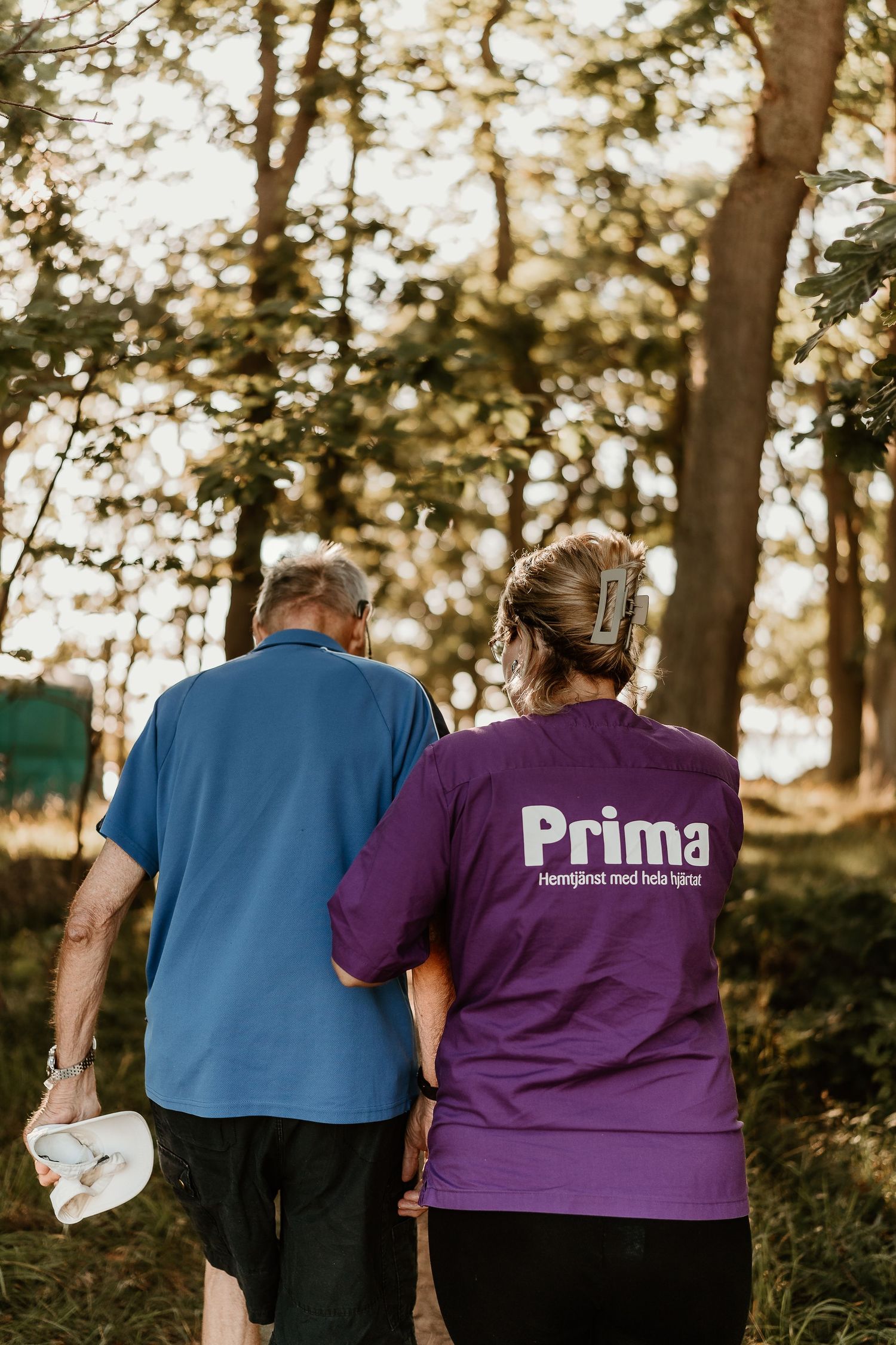 A caregiver in a Prima uniform walks with an elderly person through a sunlit wooded path.