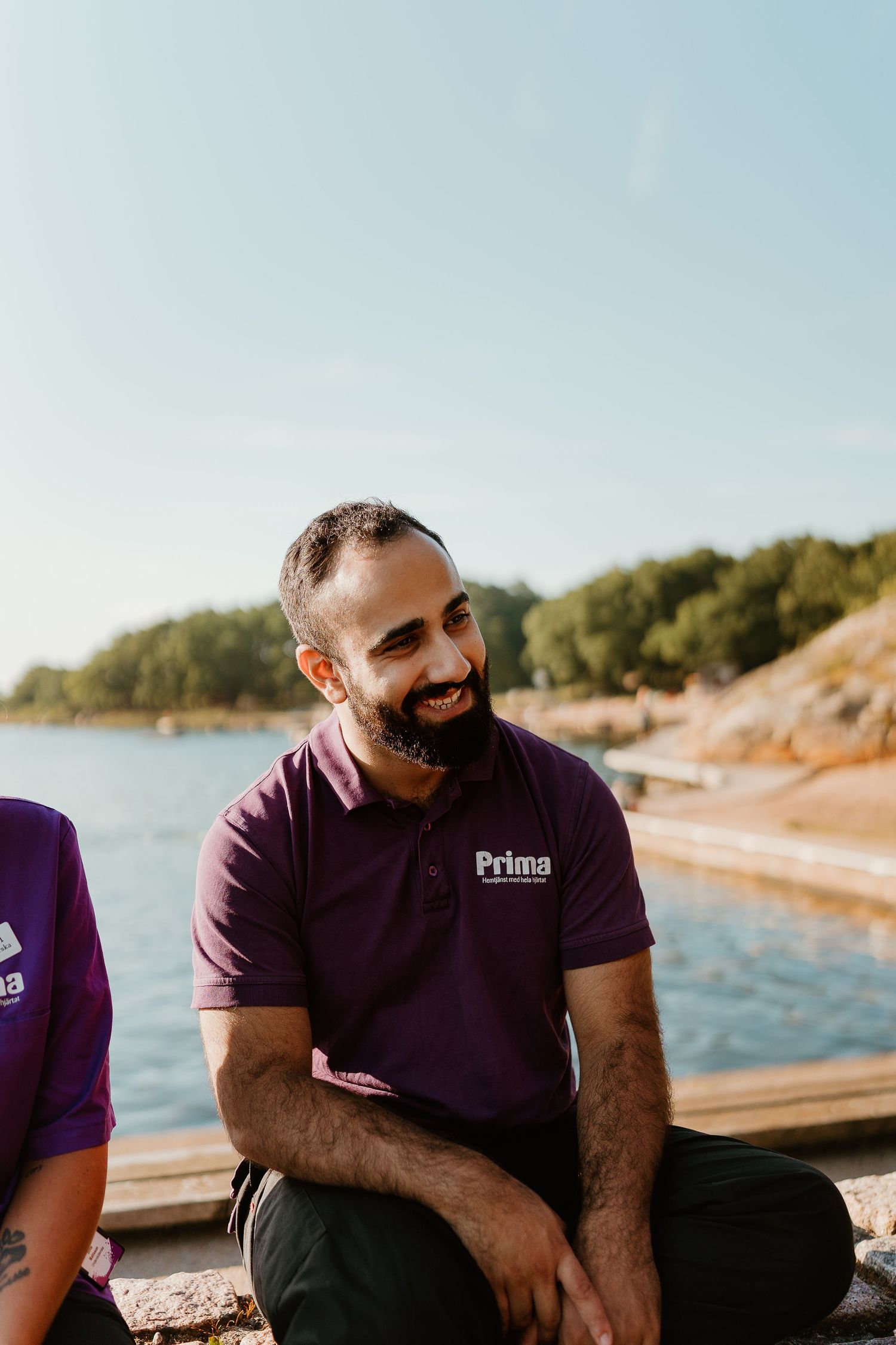 Healthcare worker in purple Prima uniform smiling while seated by waterfront.