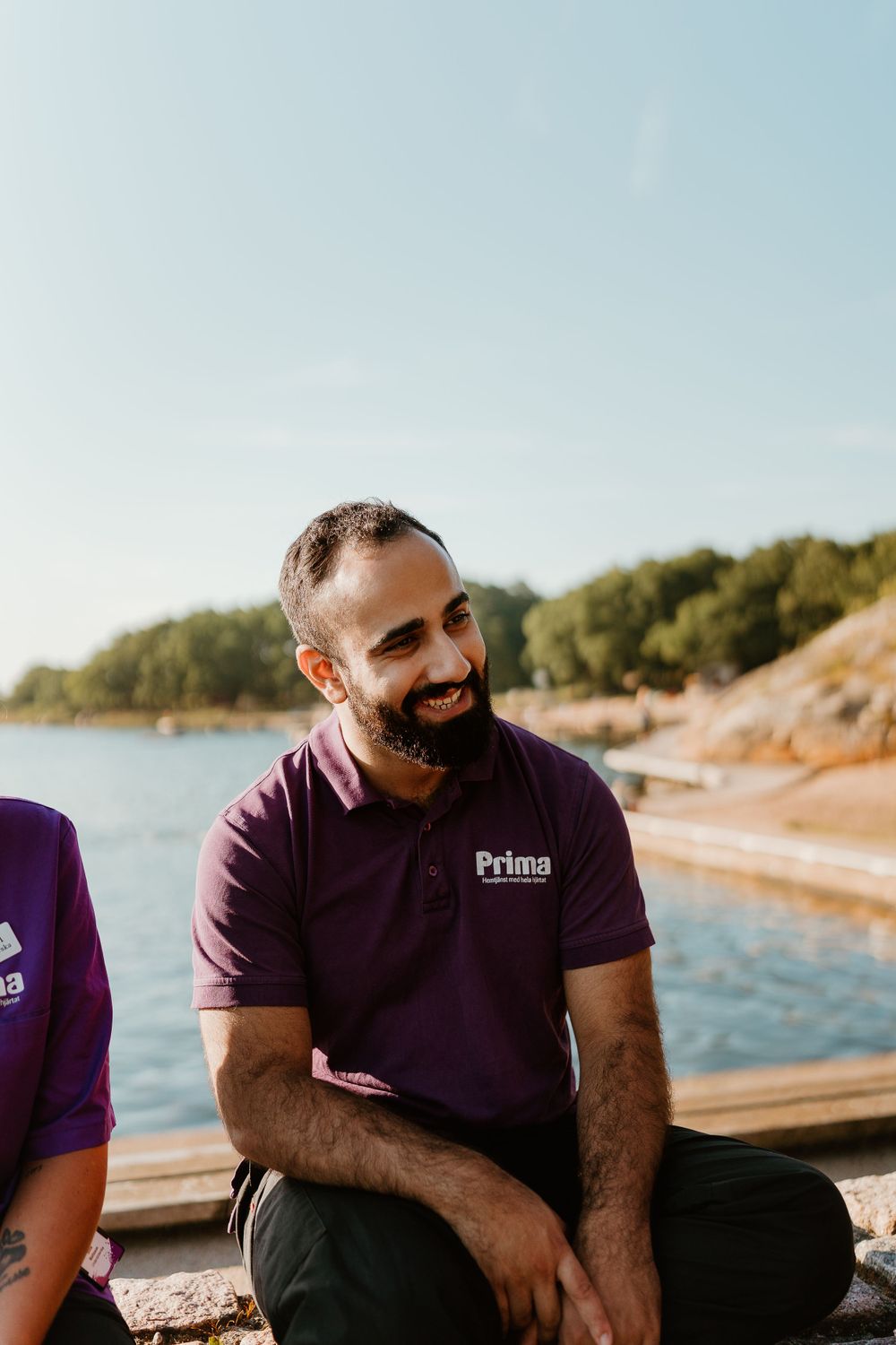 Healthcare worker in purple Prima uniform smiling while seated by waterfront.