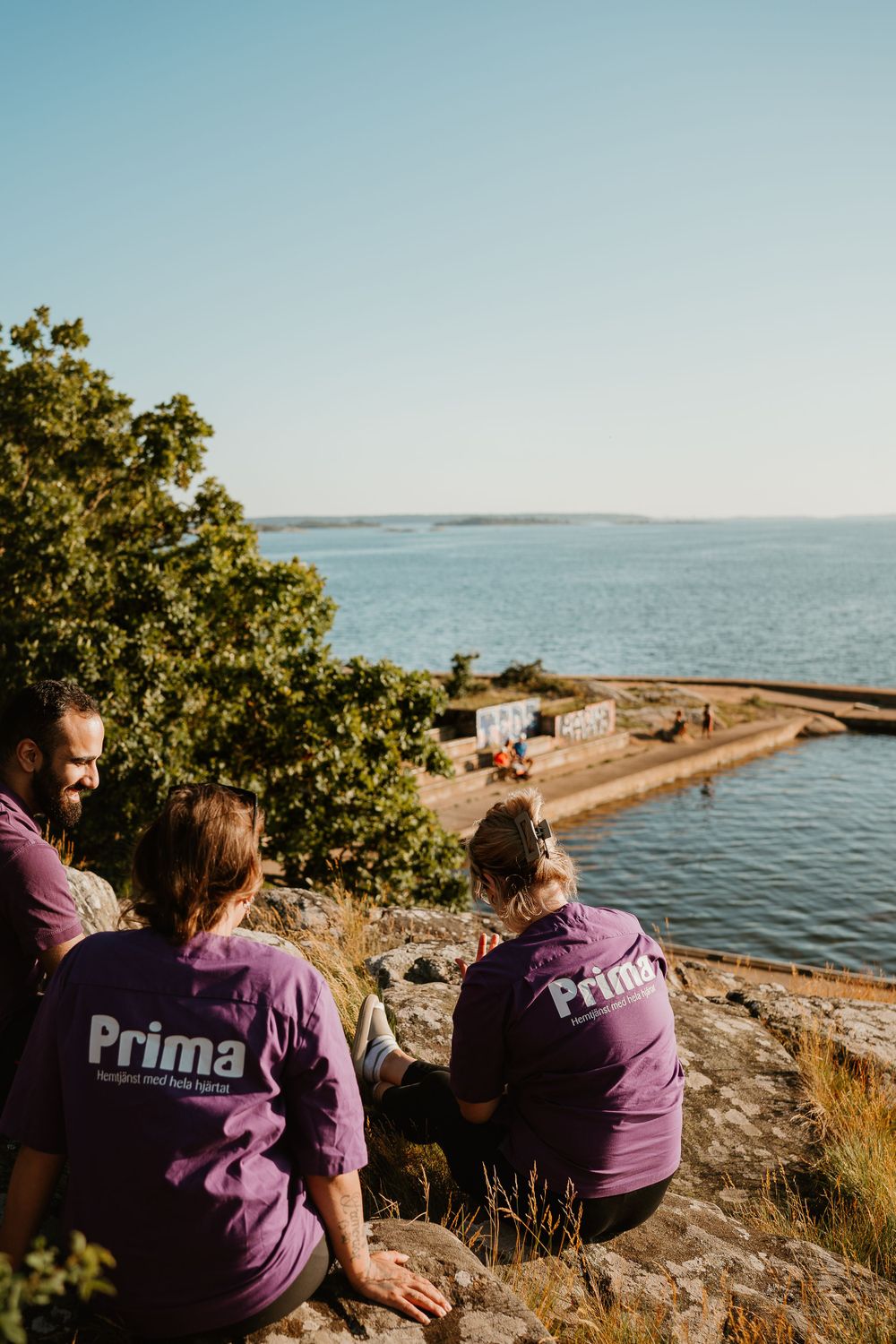 Three Prima staff members in purple shirts sitting on rocky overlook with ocean view.