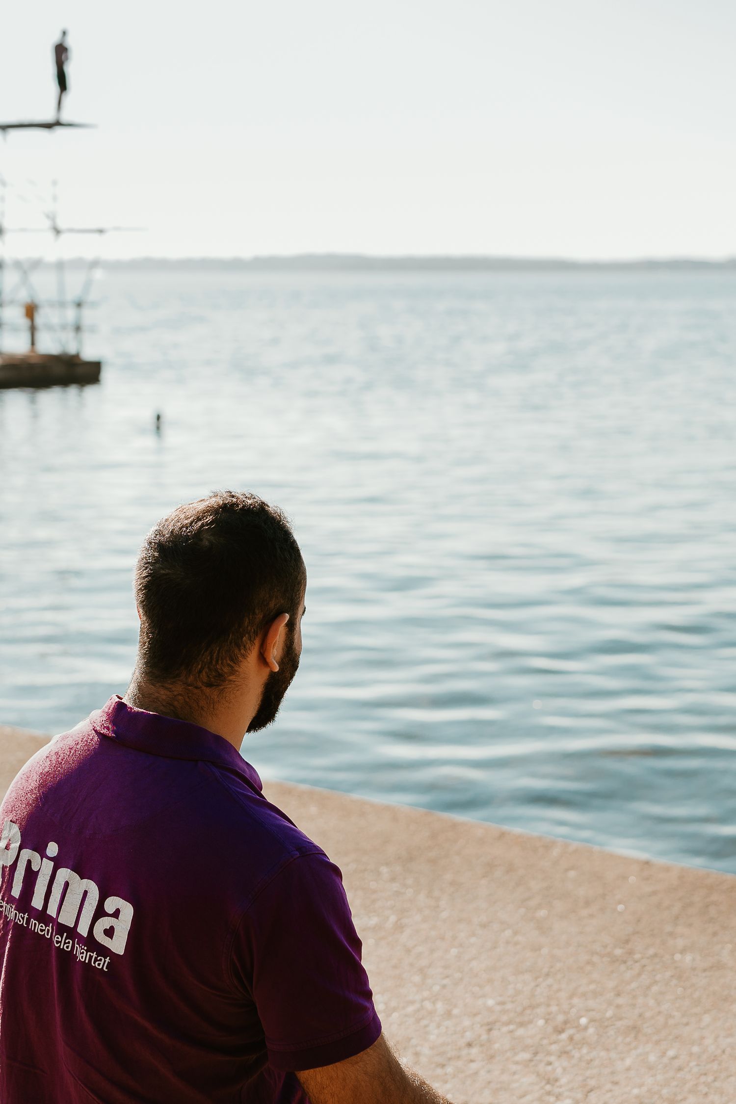A person in a purple Prima shirt sits by the seaside looking at a diving platform.