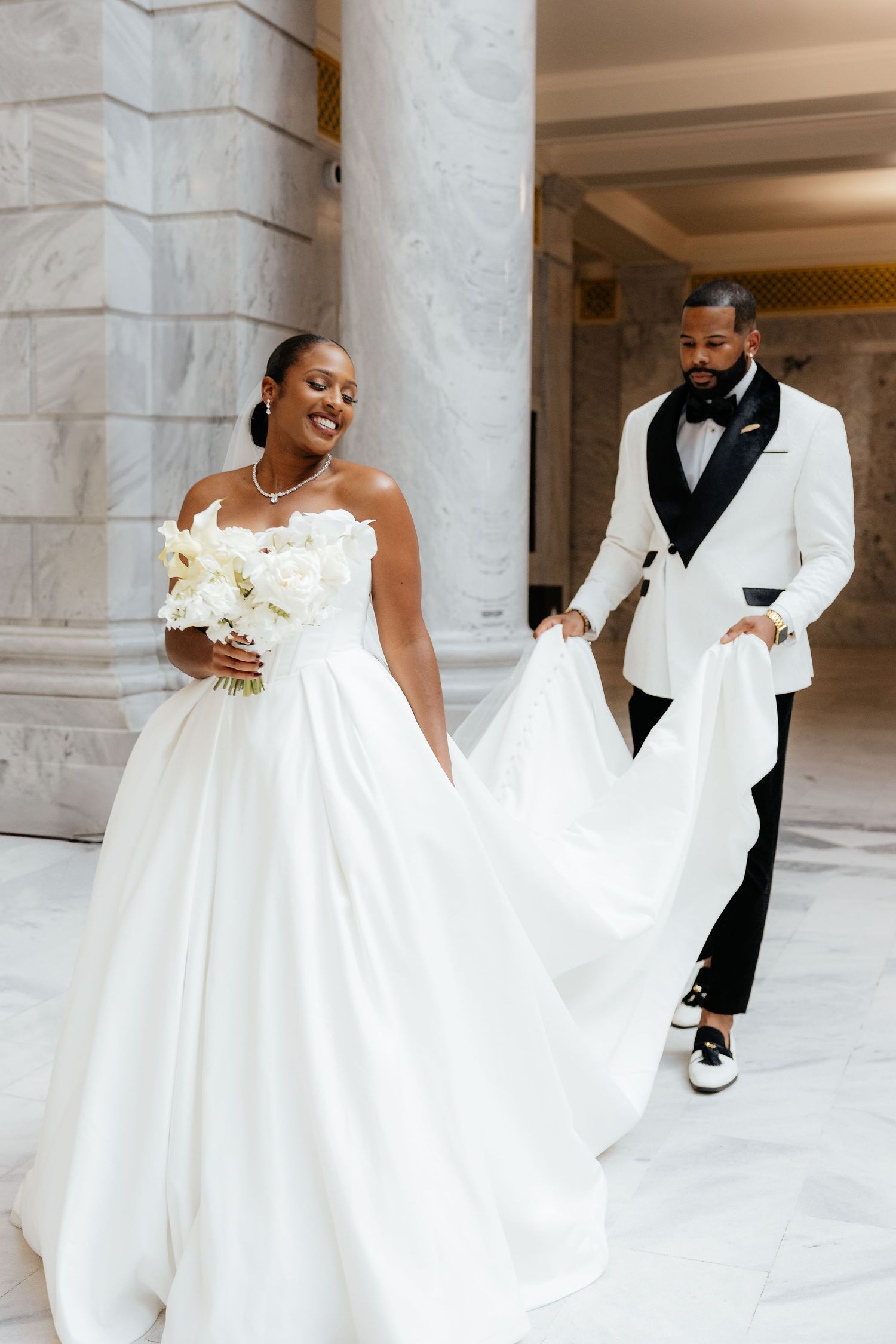 Groom in white tuxedo carries bride's white ballgown train beside marble columns at wedding.