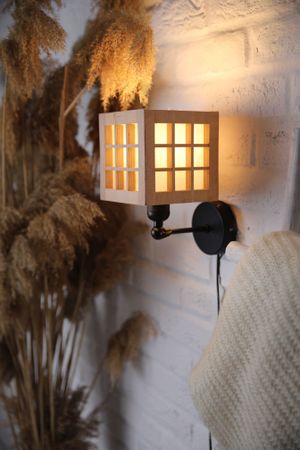 Wall-mounted wooden lattice sconce creating warm lighting against textured white wall with pampas grass.