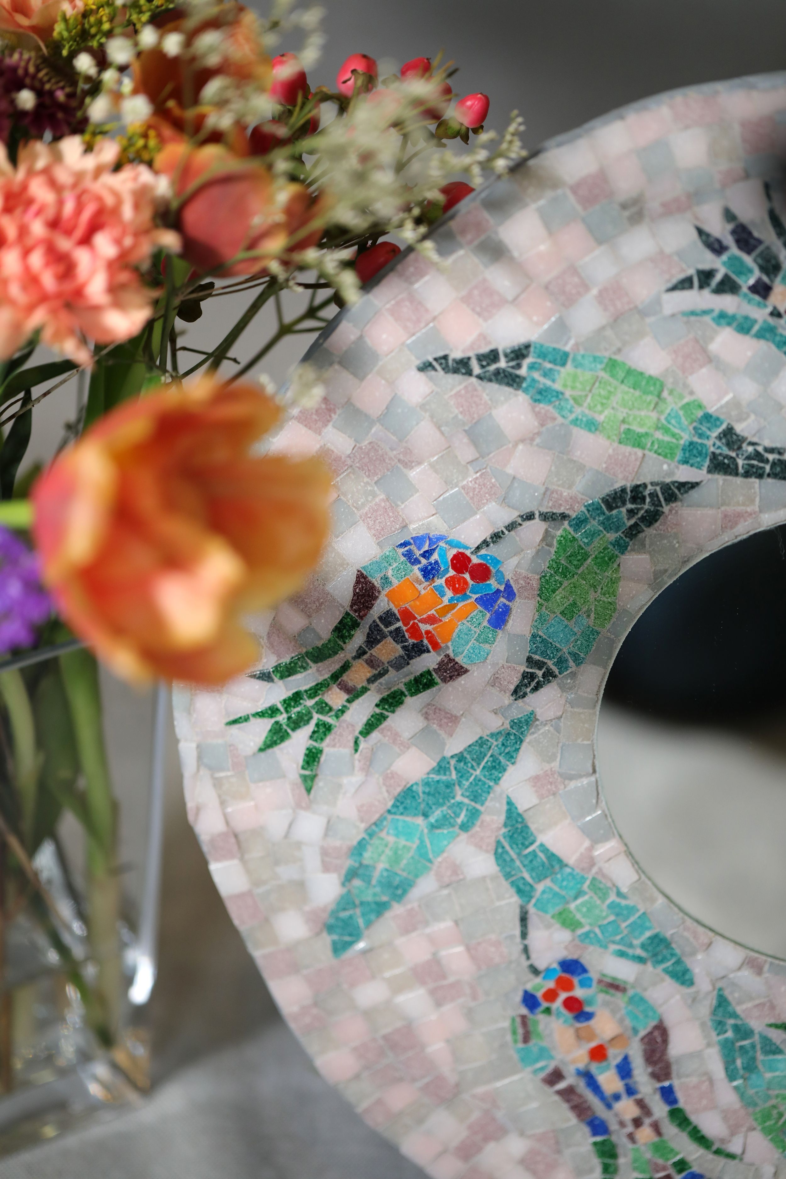 A mosaic pattern of birds and leaves in turquoise and orange tones sits beside a vase of pink and peach flowers.