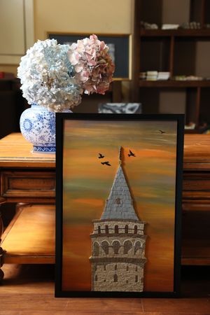 A framed artwork of Galata Tower at sunset with birds flying and blue and white vase with hydrangeas on wooden table.
