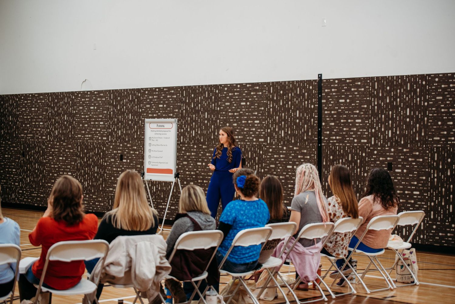 A presentation being given to an audience seated in folding chairs in front of a dark patterned wall.