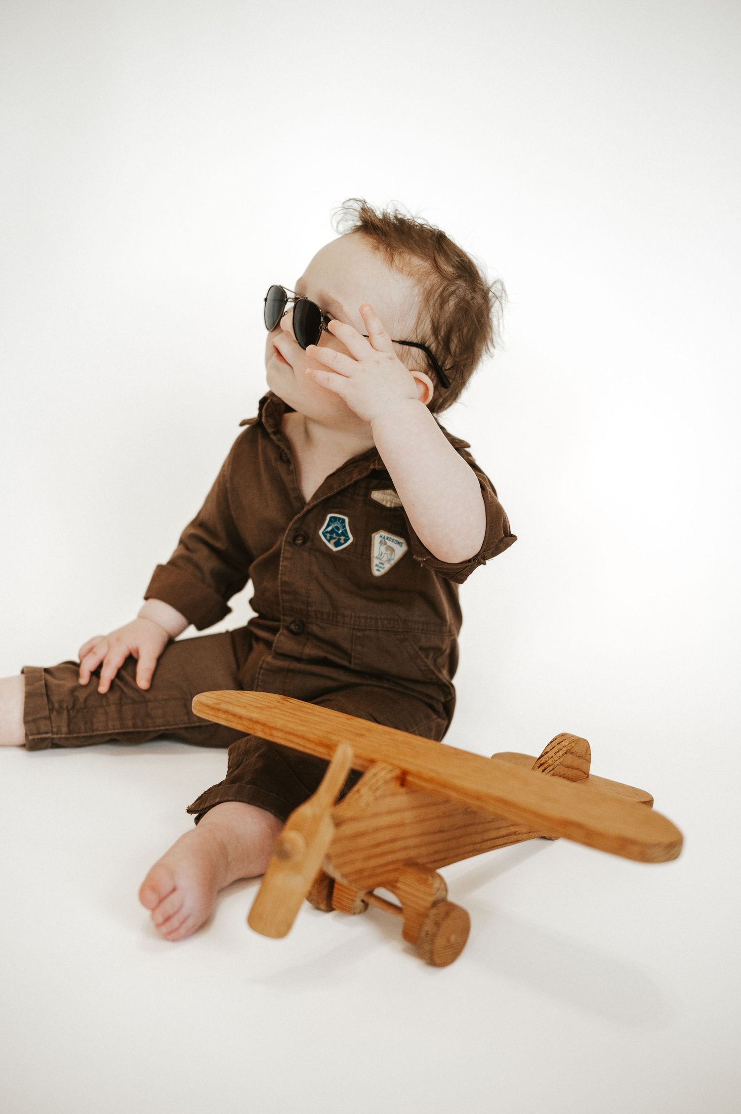 Baby in brown jumpsuit covering face while sitting with wooden toy airplane.