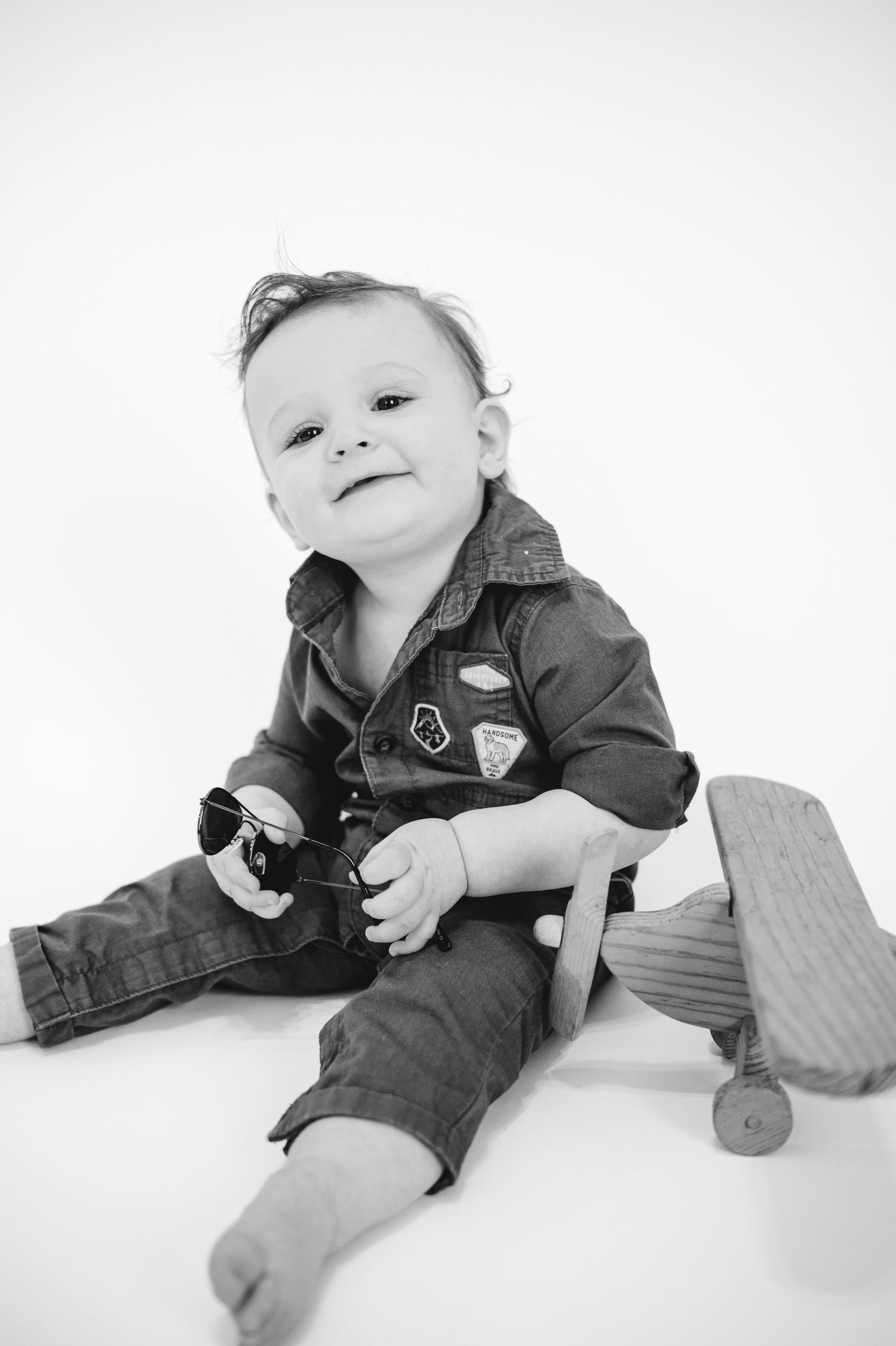 Black and white photo of infant wearing pilot outfit and playing with toy airplane.