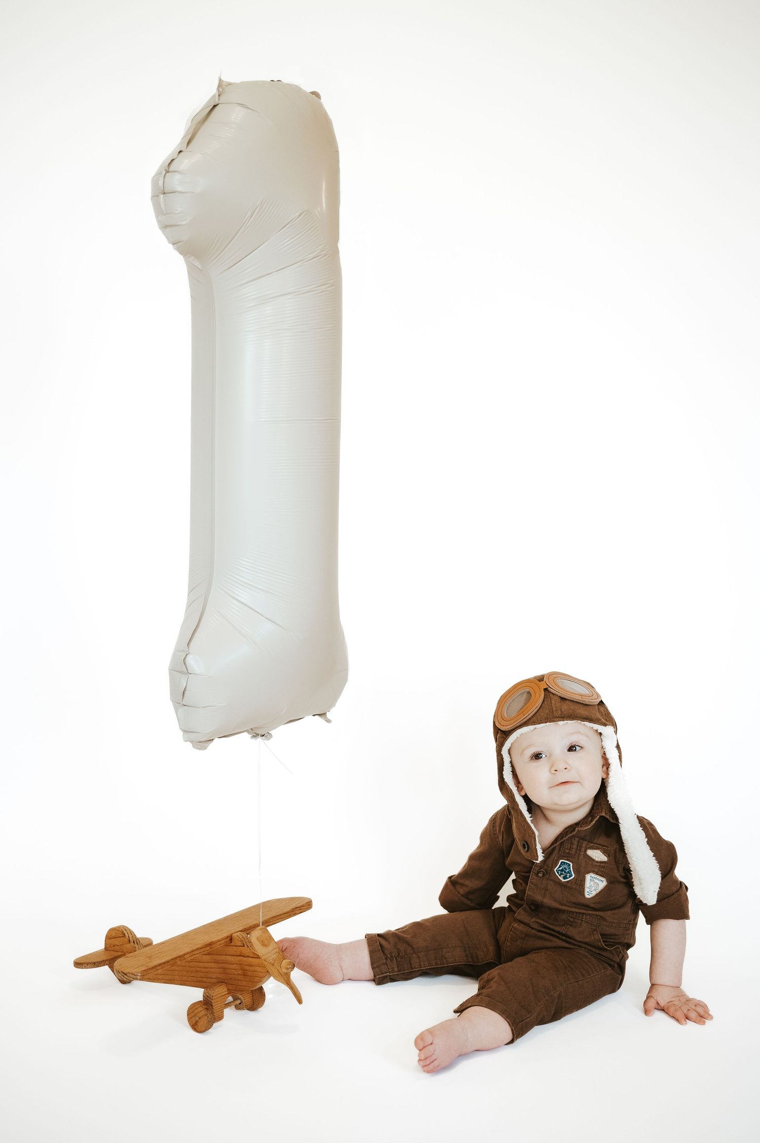 Small child in pilot outfit sitting next to large white inflatable number one and wooden toy plane.