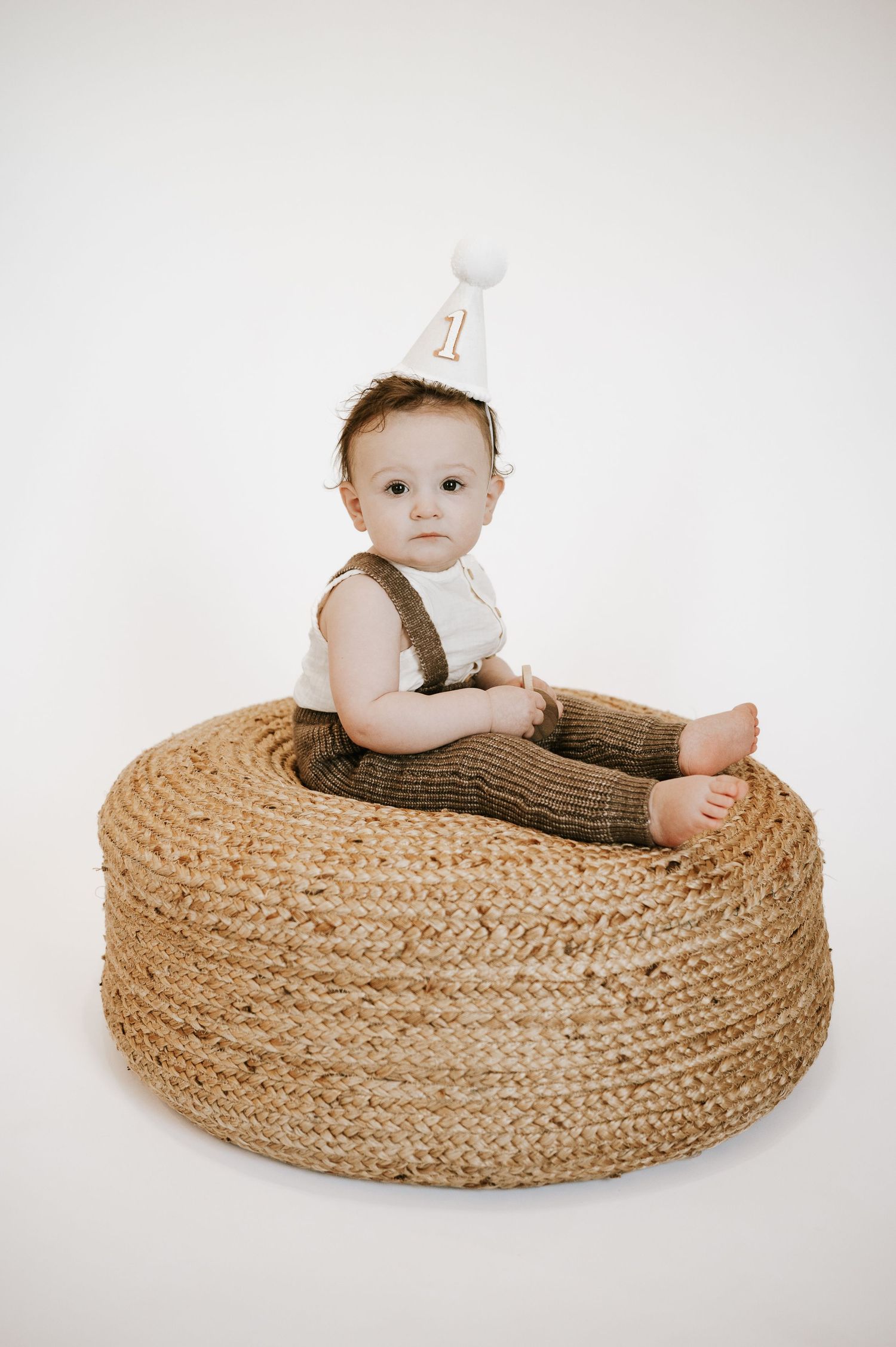 First birthday photo of baby in overalls sitting on round woven ottoman wearing party hat.