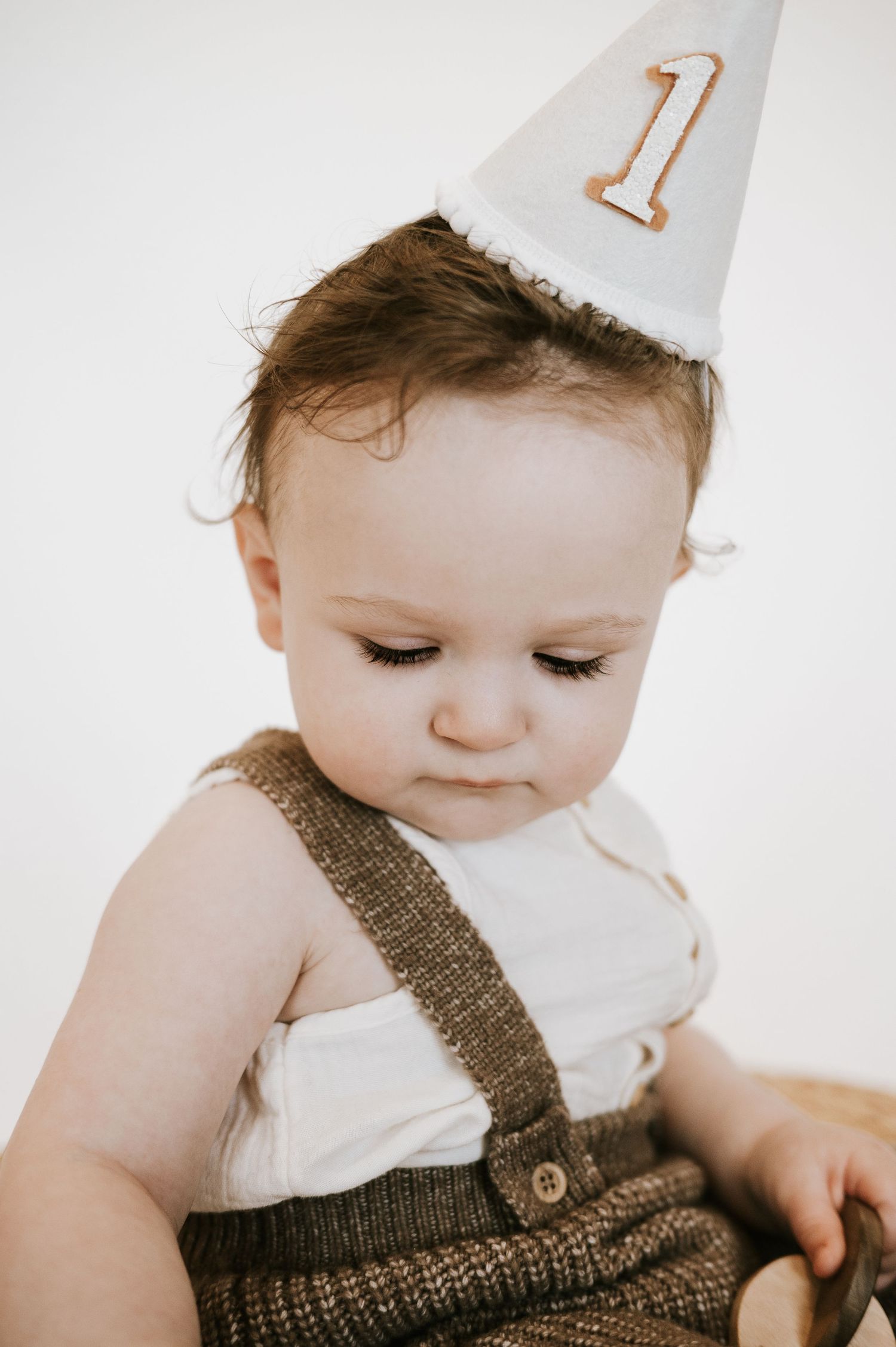 Close up of baby wearing knit overalls and white party hat with number one.
