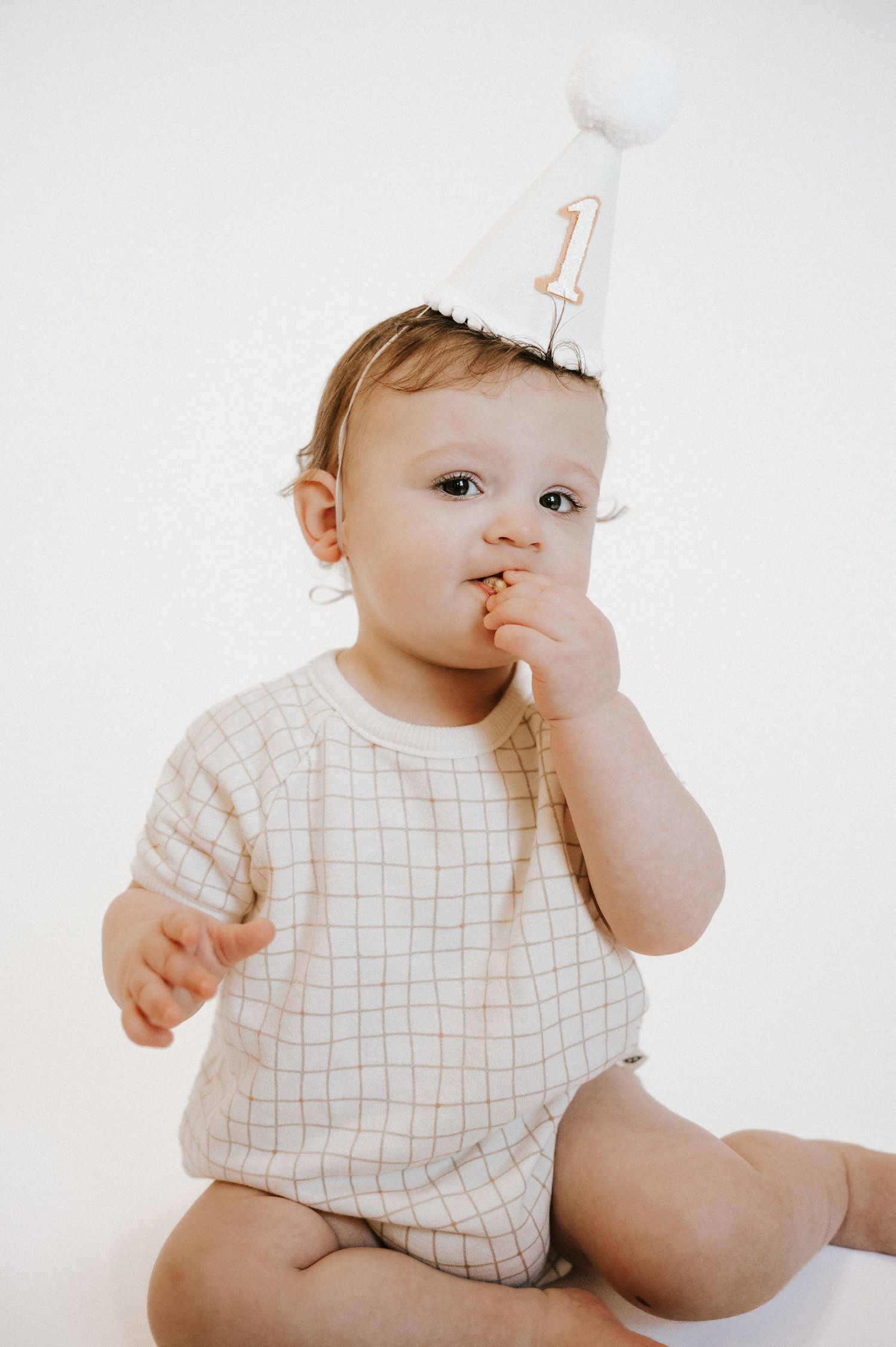 Baby wearing white grid pattern outfit and number one party hat sits looking curious on white background.