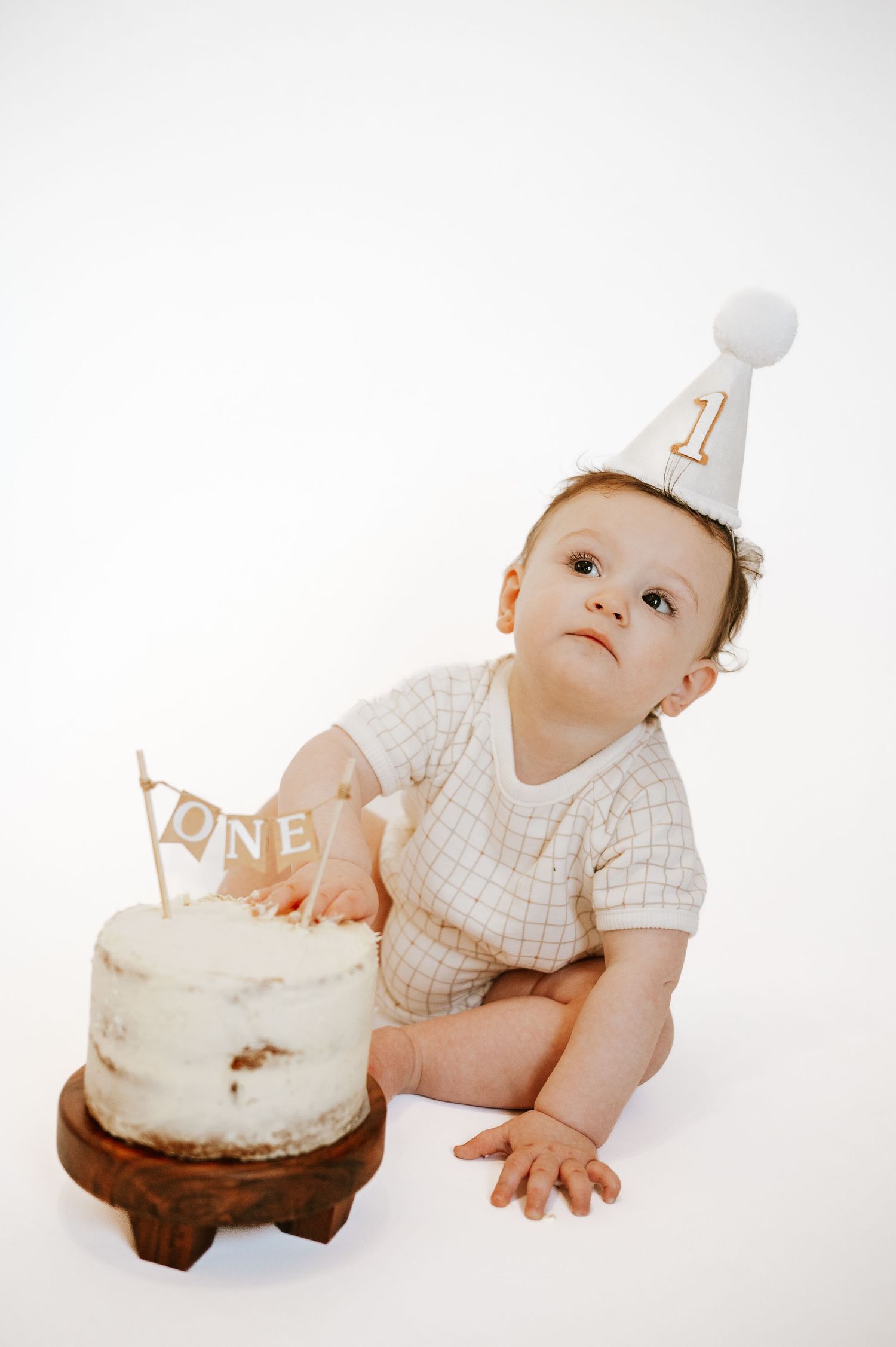 Baby in party hat reaches towards small white birthday cake on wooden stand.