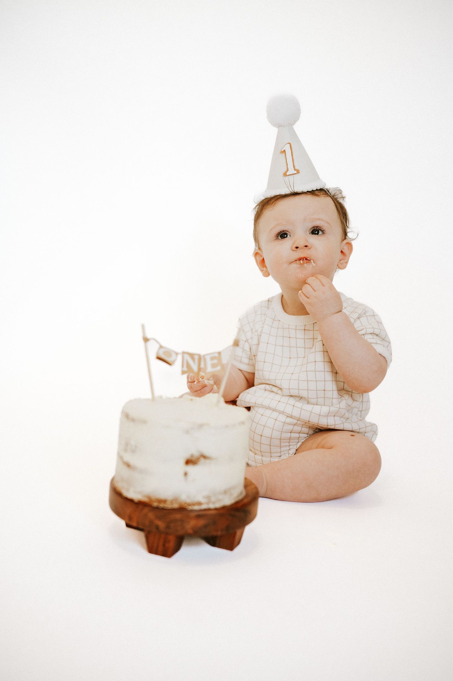 Baby in festive hat sits next to white birthday cake on wooden platform.