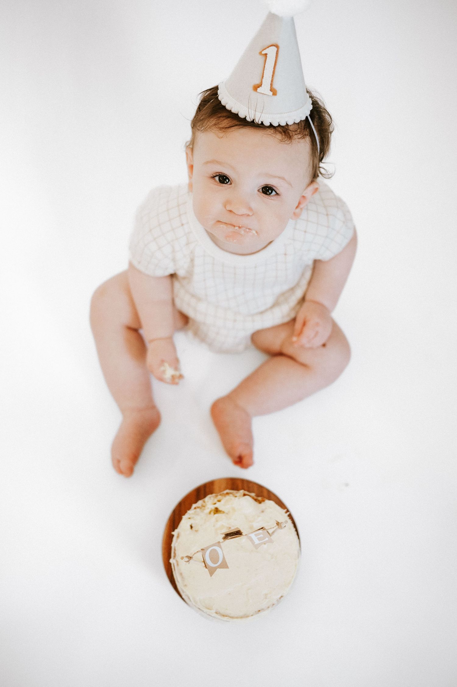 Overhead view of baby in party outfit with birthday cake during first birthday photoshoot.