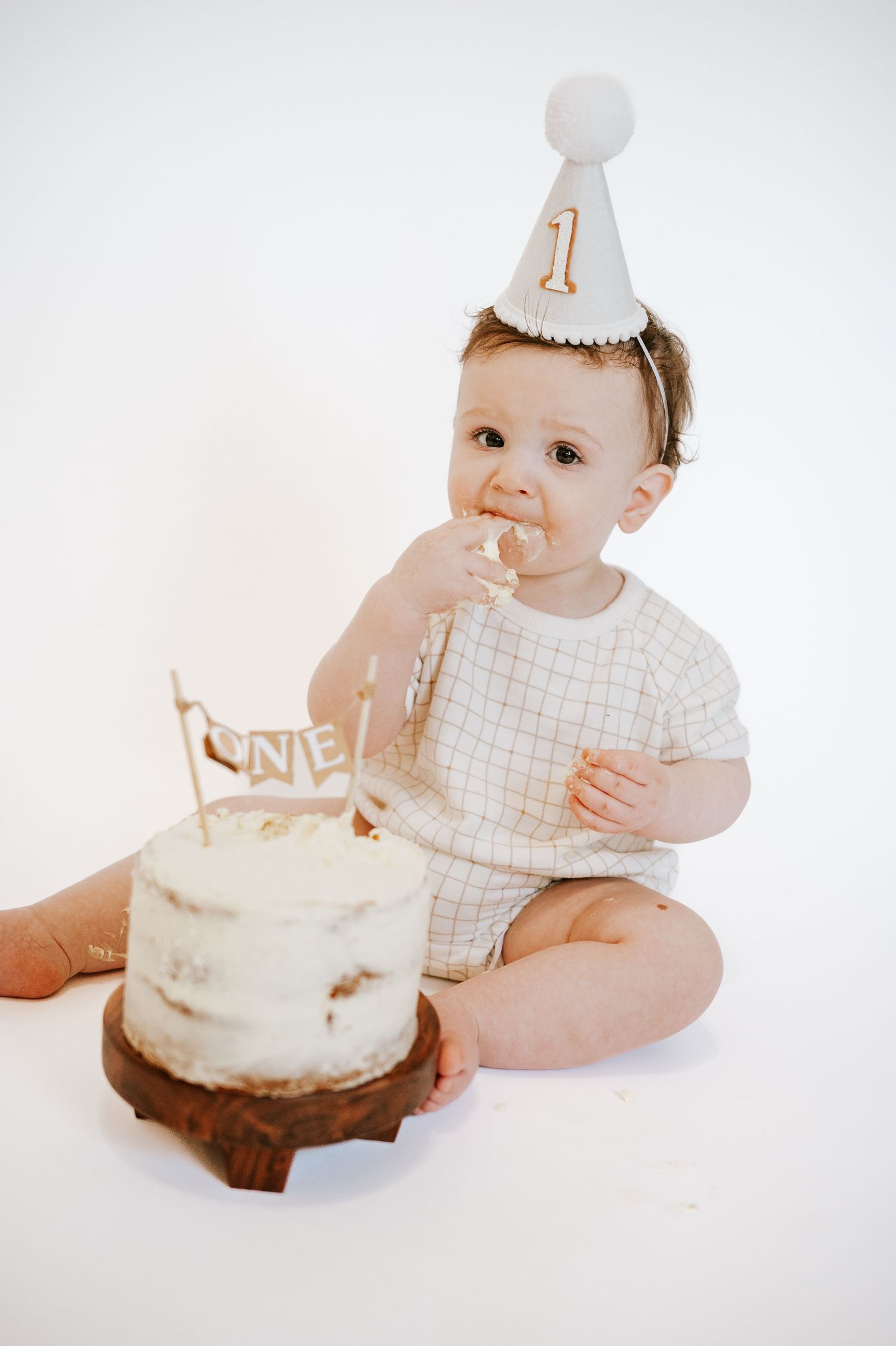 Baby in white outfit touches first birthday cake while wearing celebration hat.