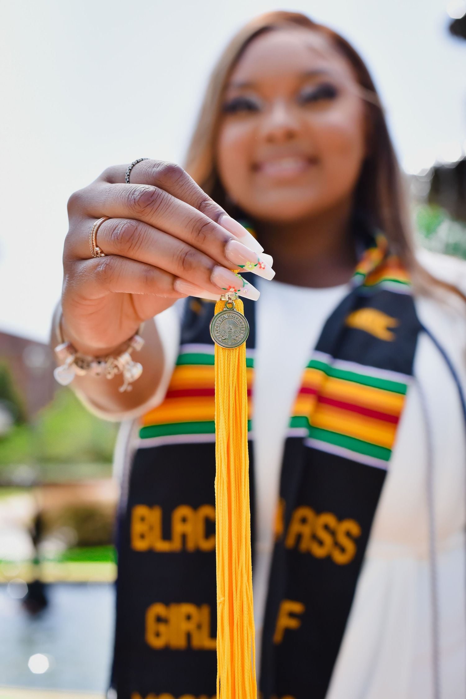 Close-up of hands holding yellow graduation tassel with cultural stole visible in background.