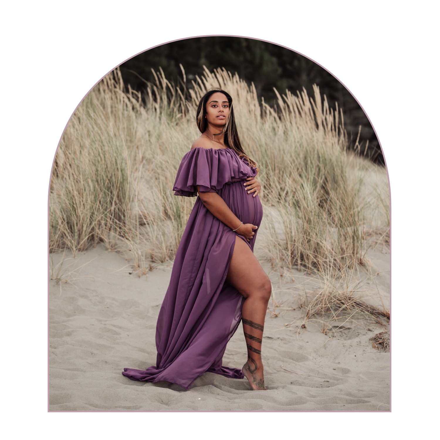 Pregnant woman in flowing purple dress poses dramatically in sand dunes during sunset photoshoot.