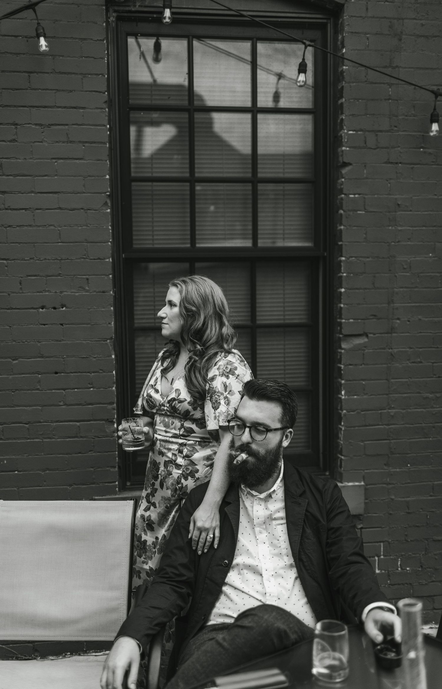 A black and white portrait of a couple relaxing against a brick wall with string lights.