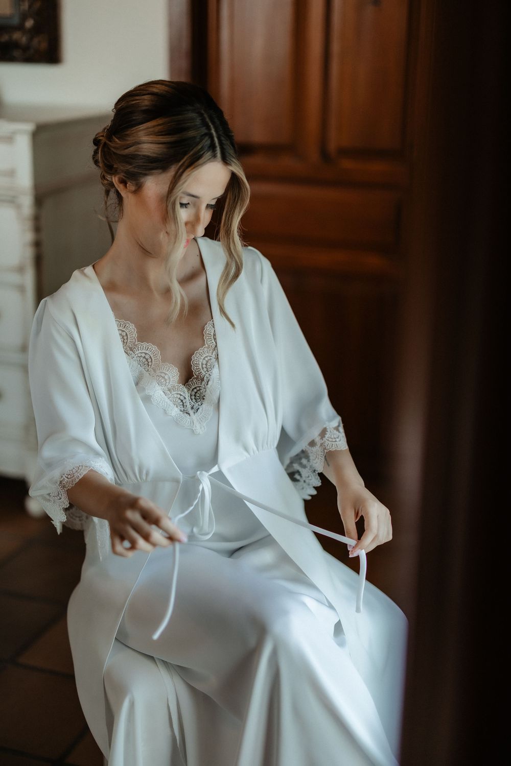 A bride in a white silk robe with lace trim prepares for her wedding day in an elegant indoor setting.
