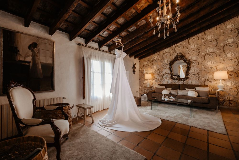 Elegant white wedding dress hanging in rustic stone room with exposed wooden beams and vintage furniture.