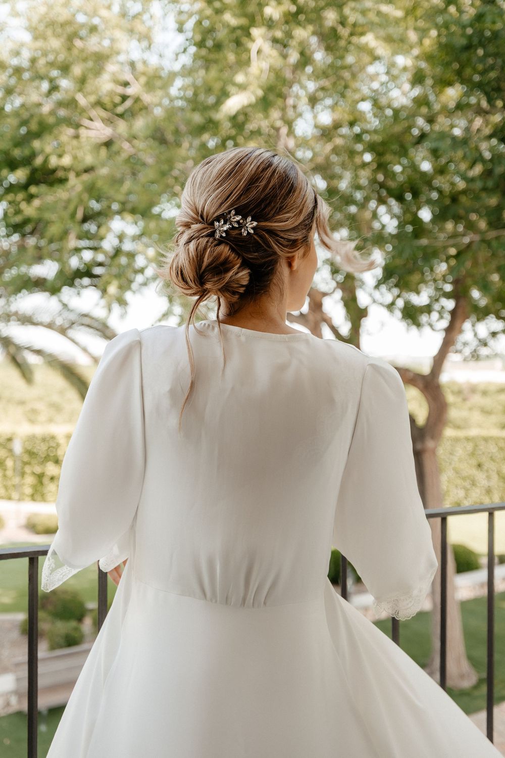 A bride in a flowing white wedding dress stands on a balcony with trees in the background.