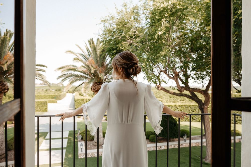 A person in a flowing white dress stands on a balcony overlooking a tropical garden with palm trees at sunset.