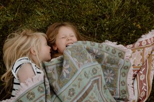 Two children lying on grass under a patterned blanket sharing a playful moment.