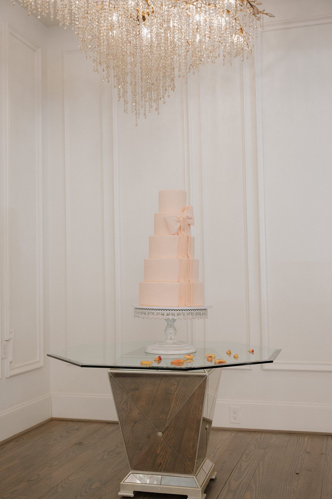 Elegant blush pink wedding cake displayed on mirrored table beneath crystal chandelier in white room.
