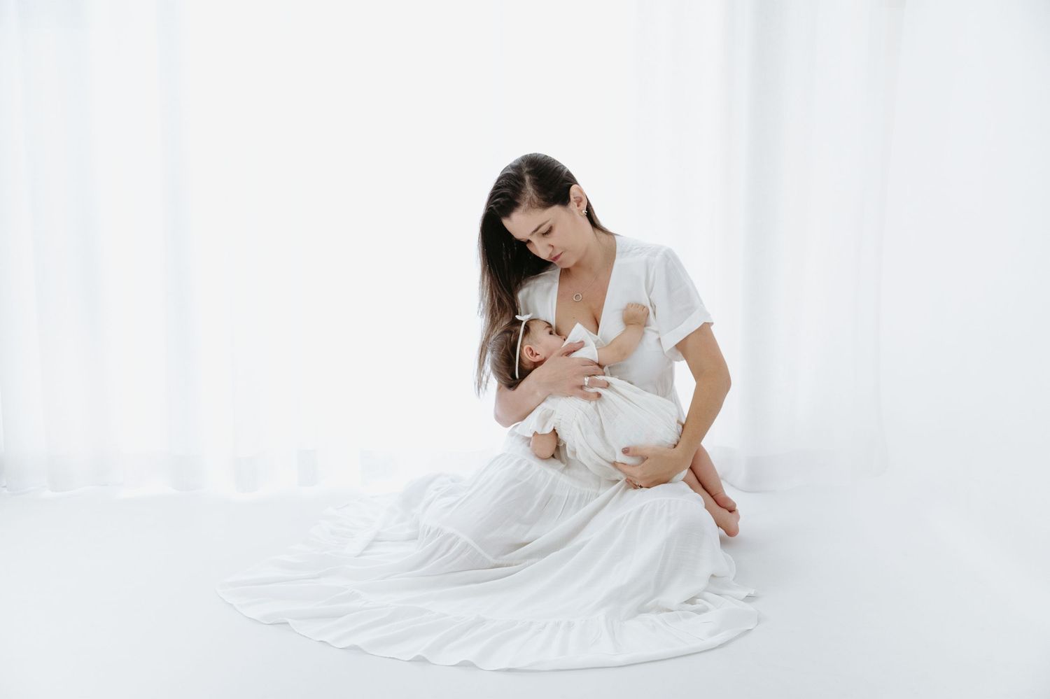 A woman sits on a white bed wearing a delicate white dress, photographed from behind.