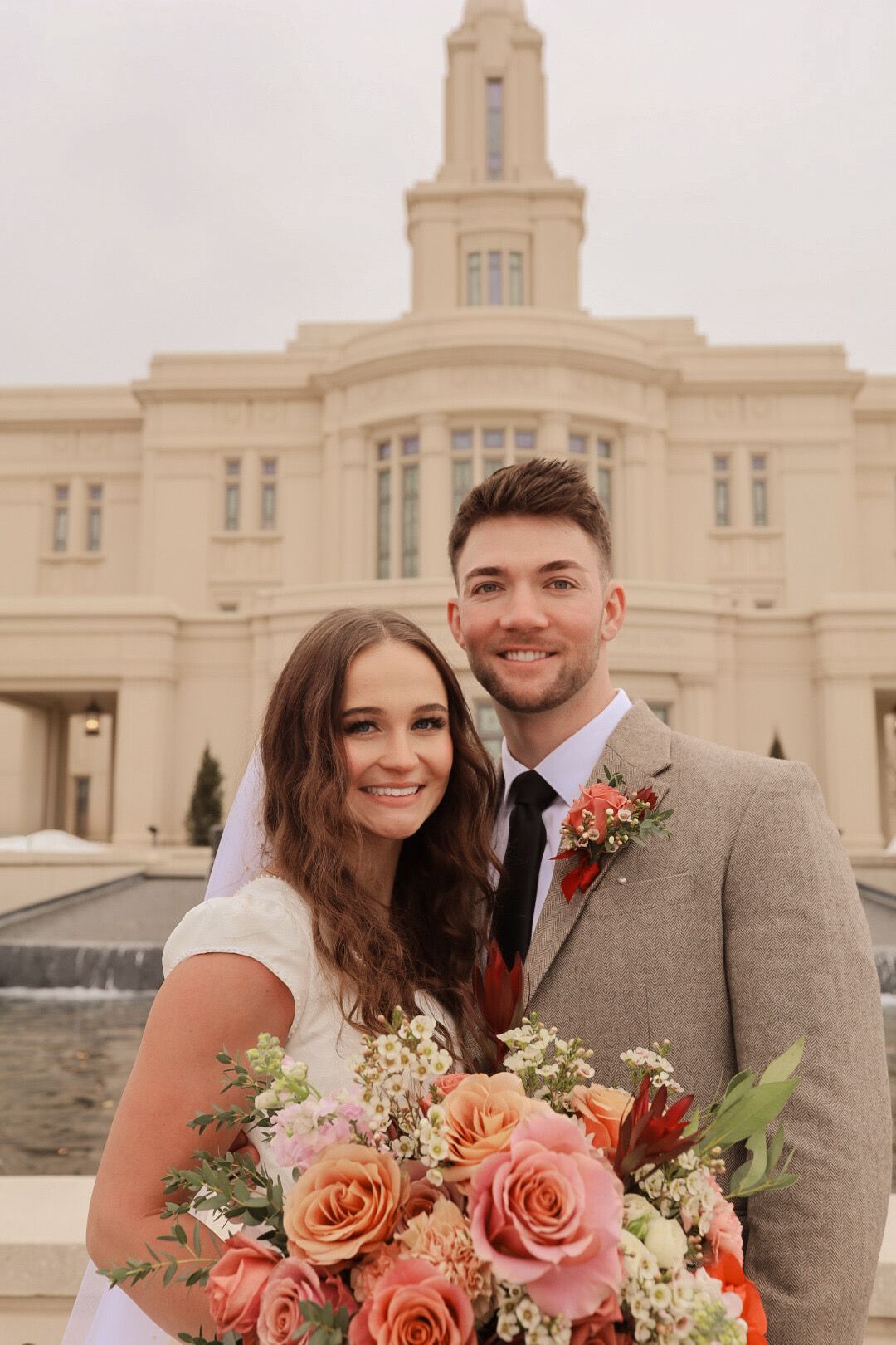 Smiling bride and groom pose with pink bouquet in front of an LDS temple on their wedding day.