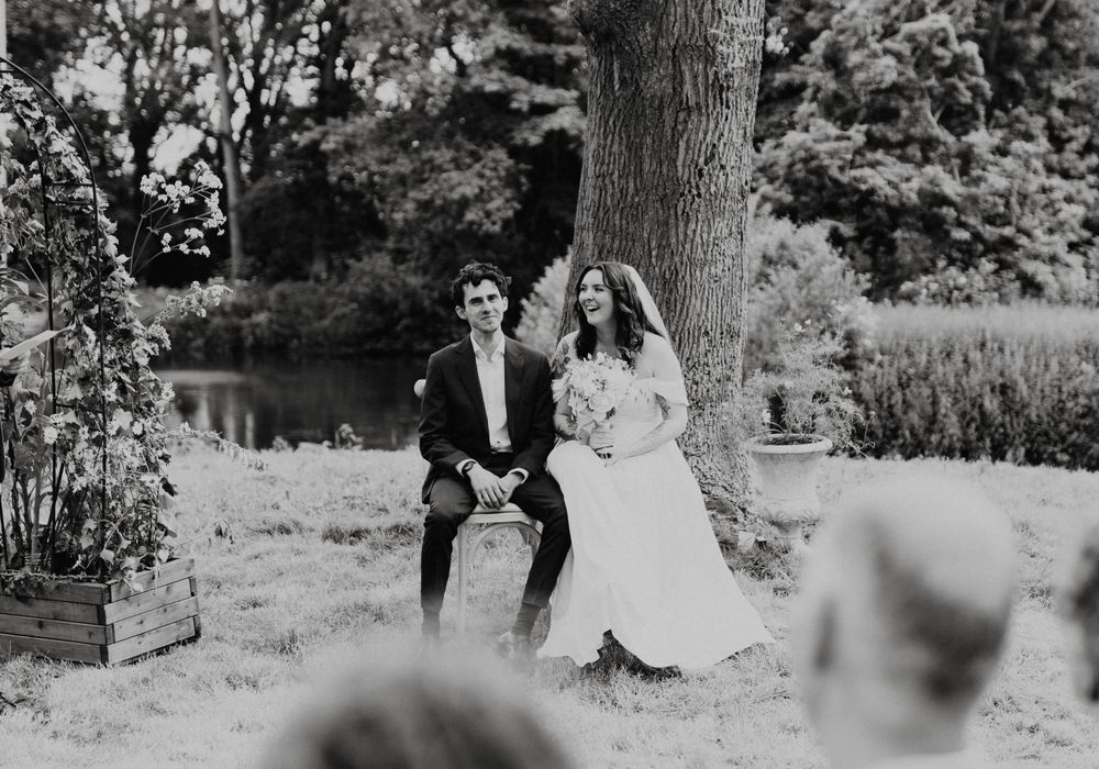 Un couple de mariés rit assis sous un arbre près d'un étang dans un moment romantique en noir et blanc.