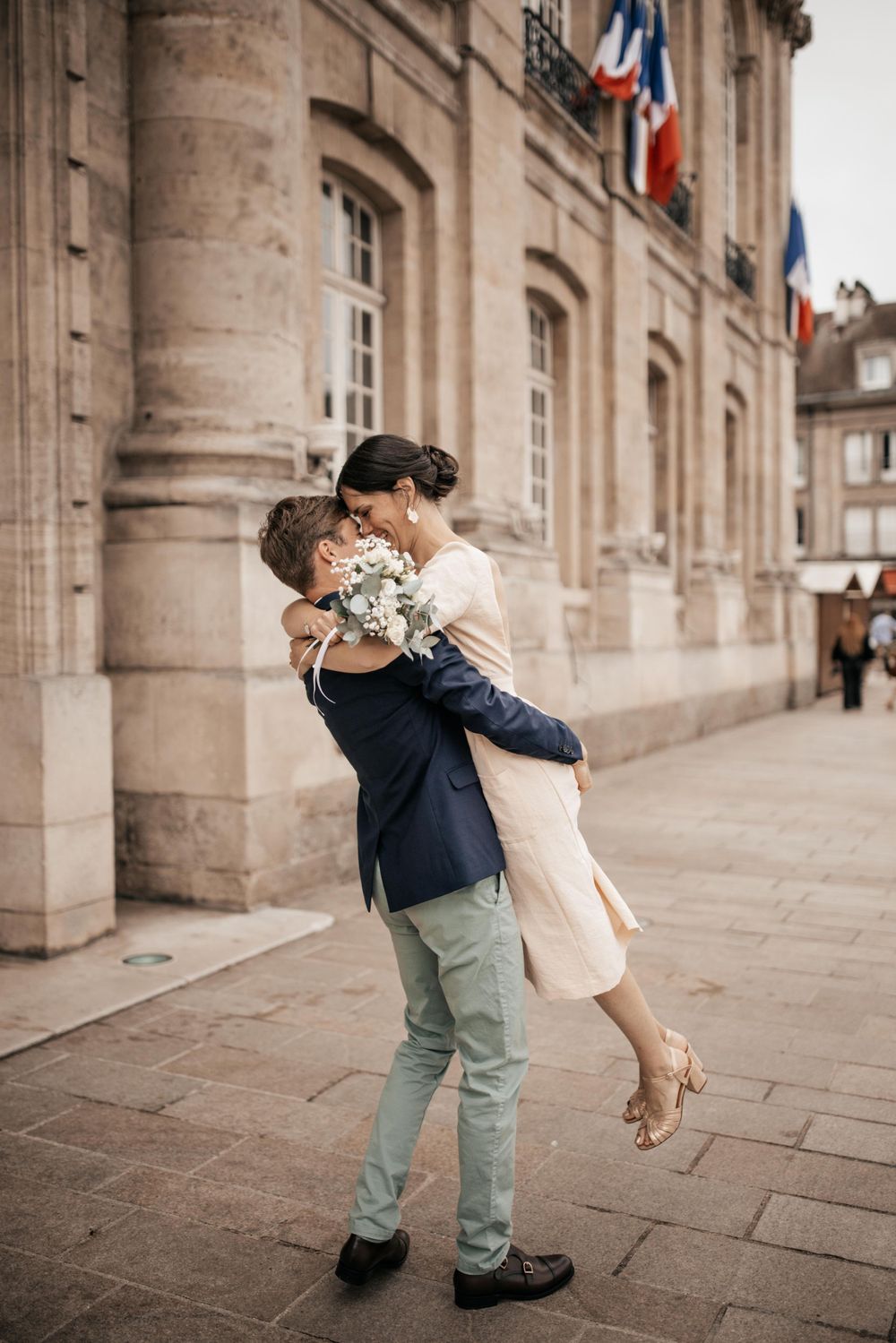 A romantic embrace between newlyweds in front of a historic French building with flags.