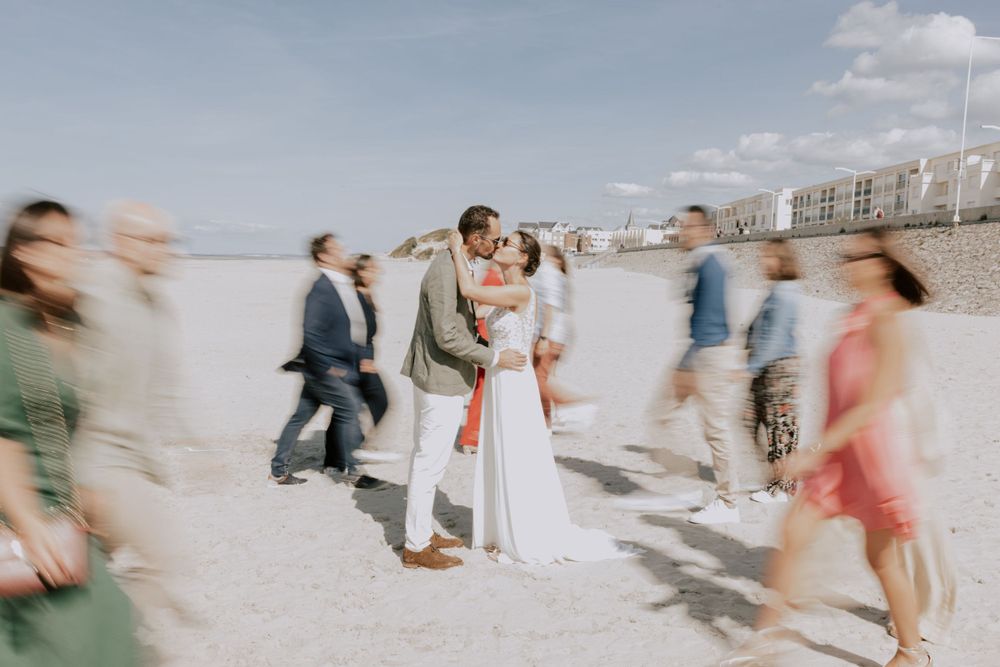 Scène de mariage floue sur la plage avec un couple s'embrassant tandis que les invités passent.