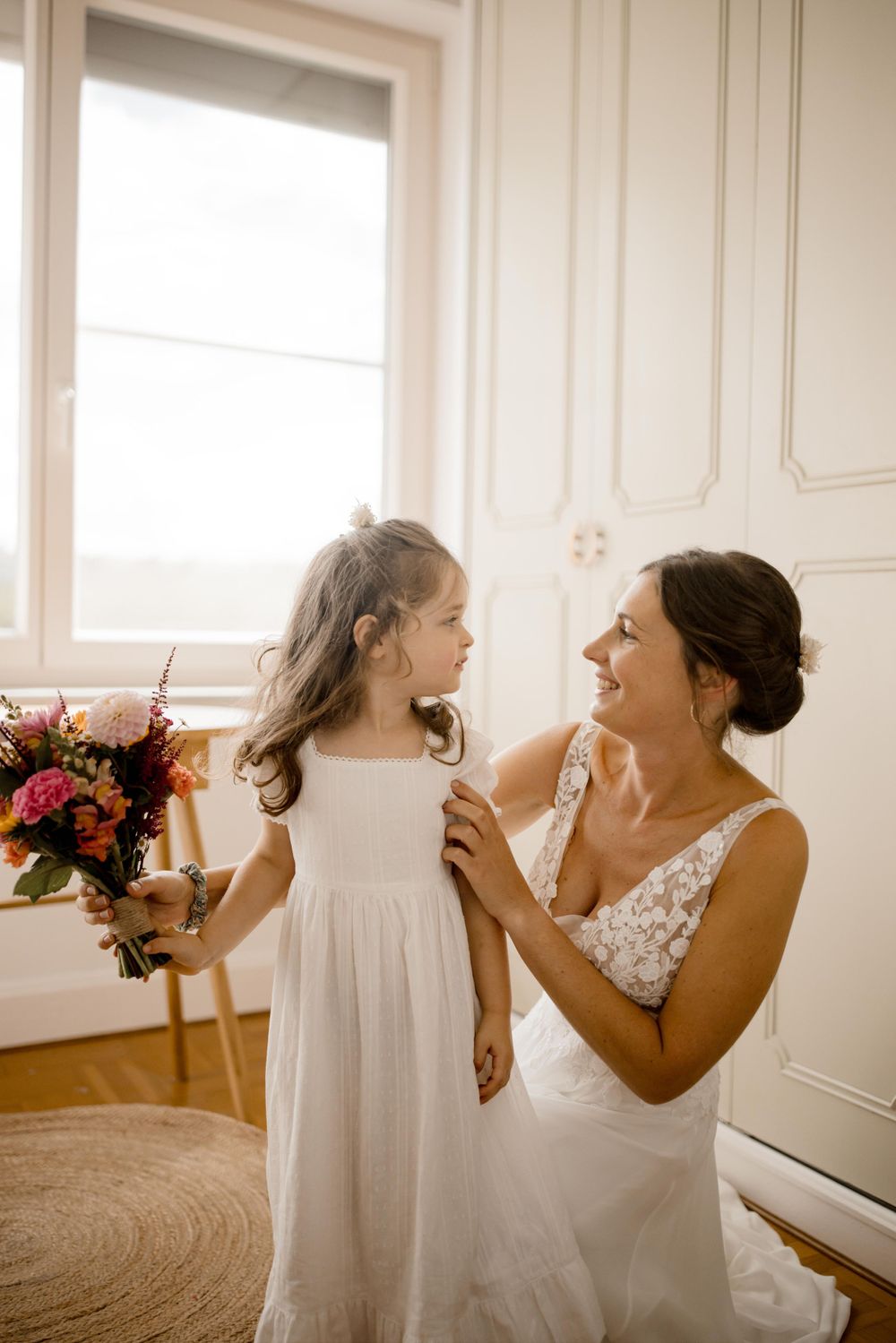 Moment de tendresse entre la mariée en robe de dentelle et la jeune fille en robe blanche devant une fenêtre lumineuse.