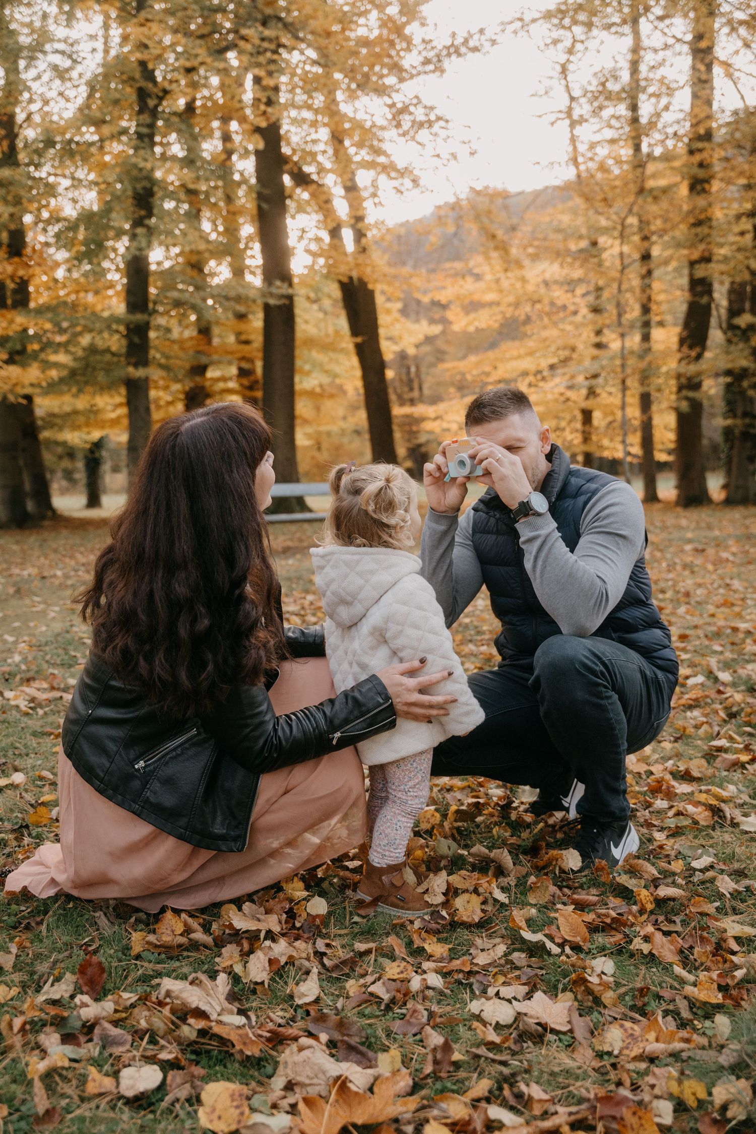 A family enjoys a tender moment together on fallen autumn leaves in a golden forest setting.