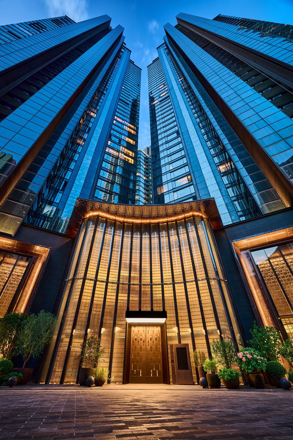 Luxury skyscraper entrance with golden facade, glass towers, and lush potted plants under a blue evening sky.