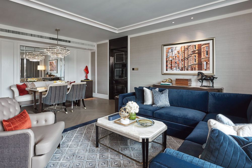 Elegant living room with blue velvet sofa, gray armchairs, geometric rug, and dining area with chandelier.