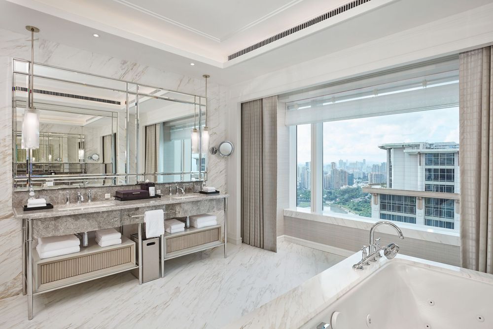 Luxurious marble hotel bathroom with dual vanity, large mirror, soaking tub, and panoramic city views through a wide window.